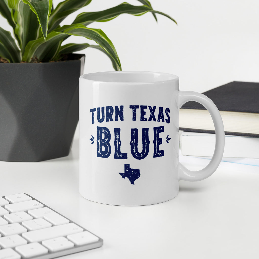 White ceramic coffee mug featuring a distressed blue "Turn Texas Blue" design with a Texas state silhouette, on a desktop with keyboard, book, and plant.