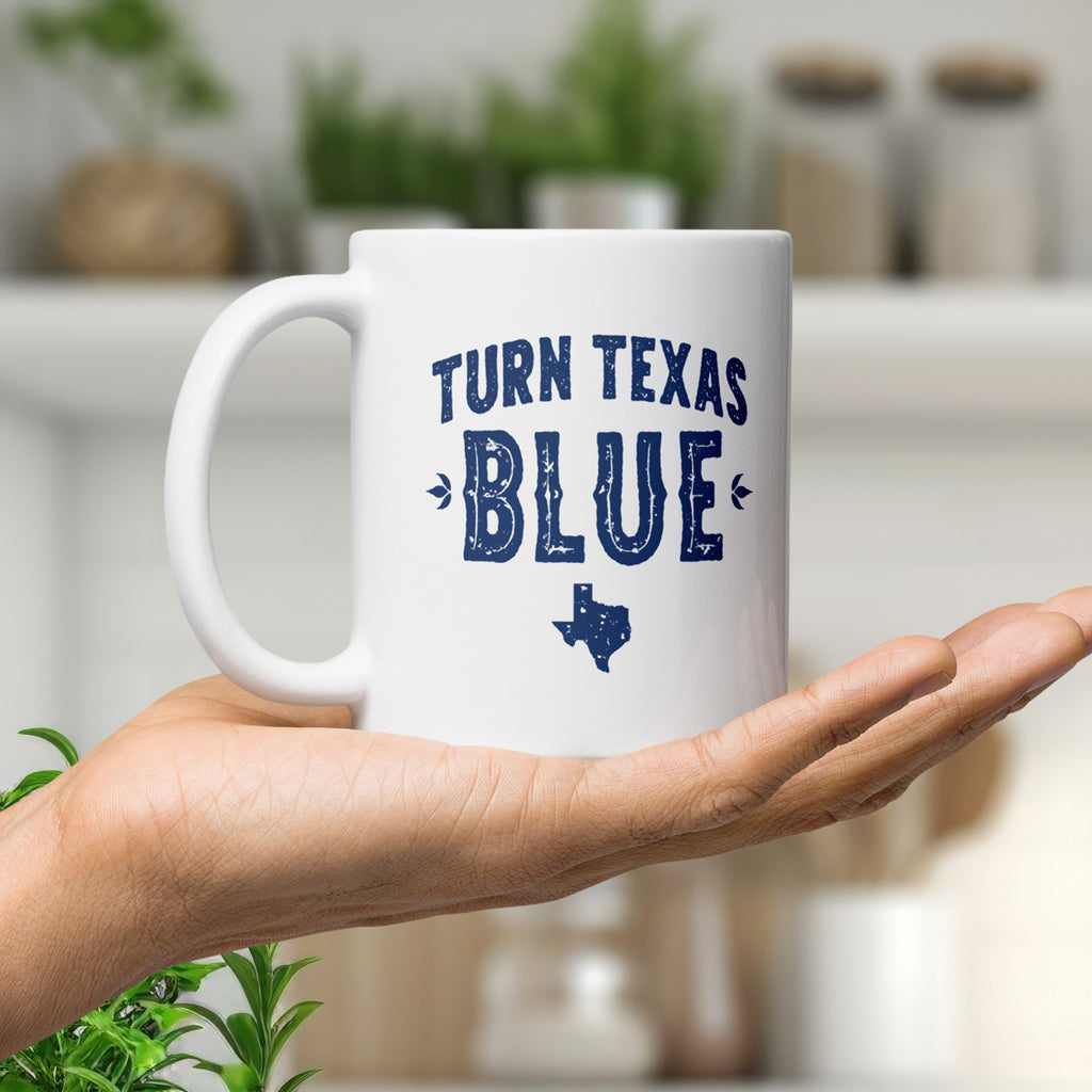 White ceramic coffee mug featuring a distressed blue "Turn Texas Blue" design with a Texas state silhouette, held in the palm of a hand in a nice kitchen.