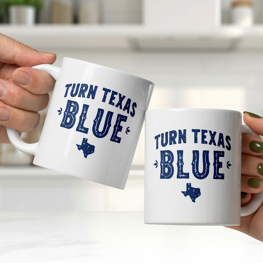 Two white ceramic coffee mugs featuring the same distressed blue "Turn Texas Blue" design with a Texas state silhouette on each side, held by two people, clinking the mugs together in a white kitchen.