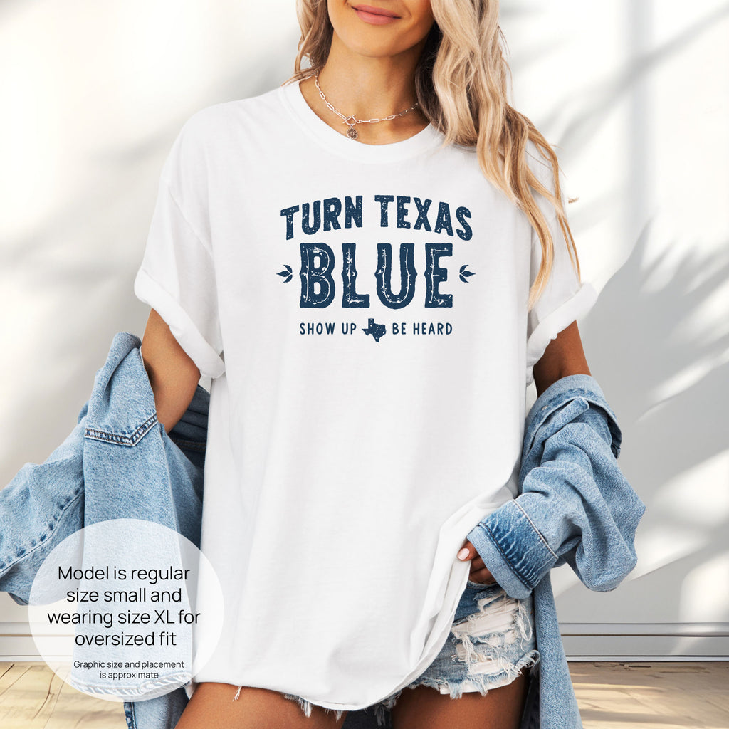 Female model wearing oversized white t-shirt featuring distressed "Turn Texas Blue" text with a small Texas icon and the phrase "Show Up Be Heard." Includes size and fit information for the model who is regular size small, and wearing a size XL shirt for an oversized fit.

