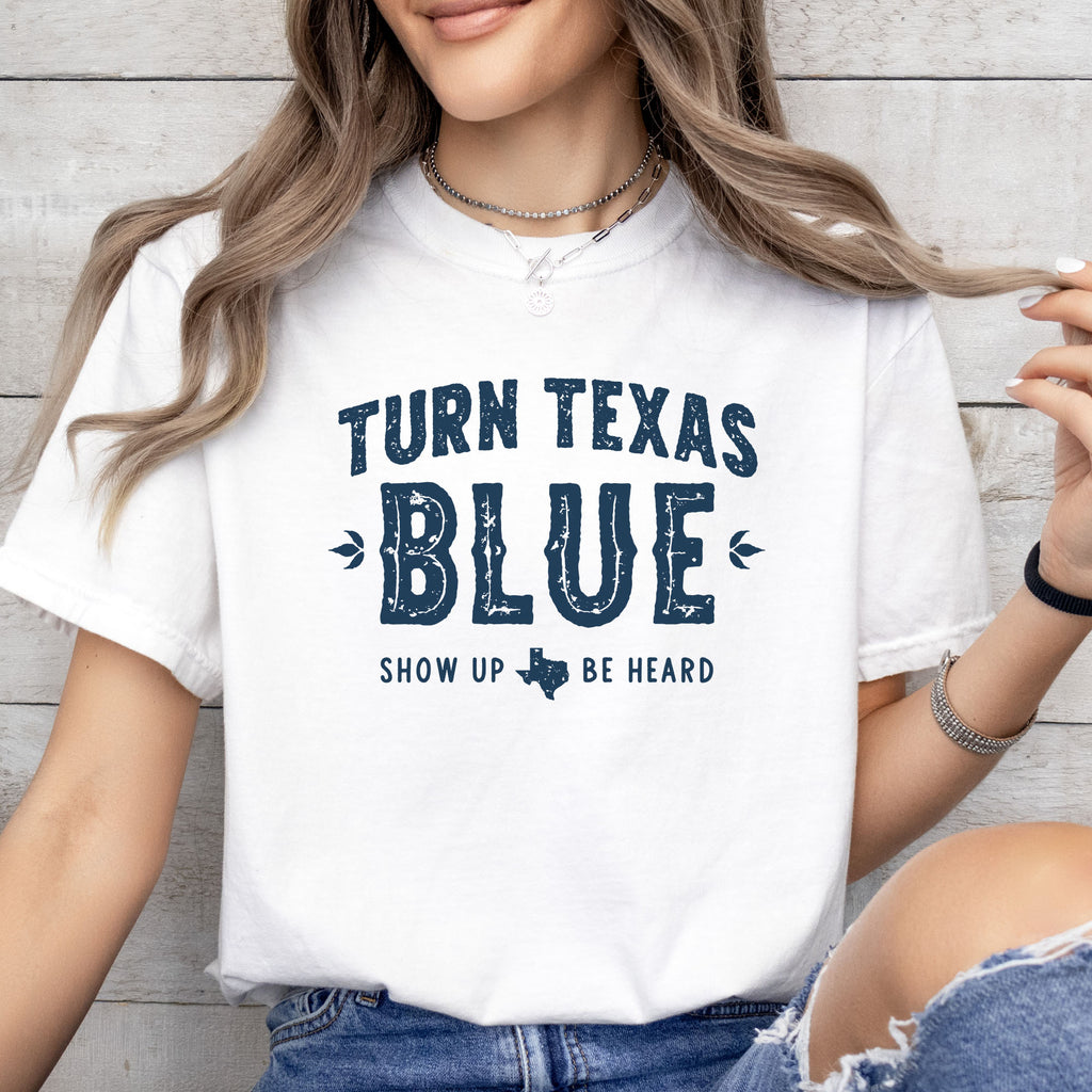 Female model wearing white t-shirt featuring distressed "Turn Texas Blue" text with a small Texas icon and the phrase "Show Up Be Heard."