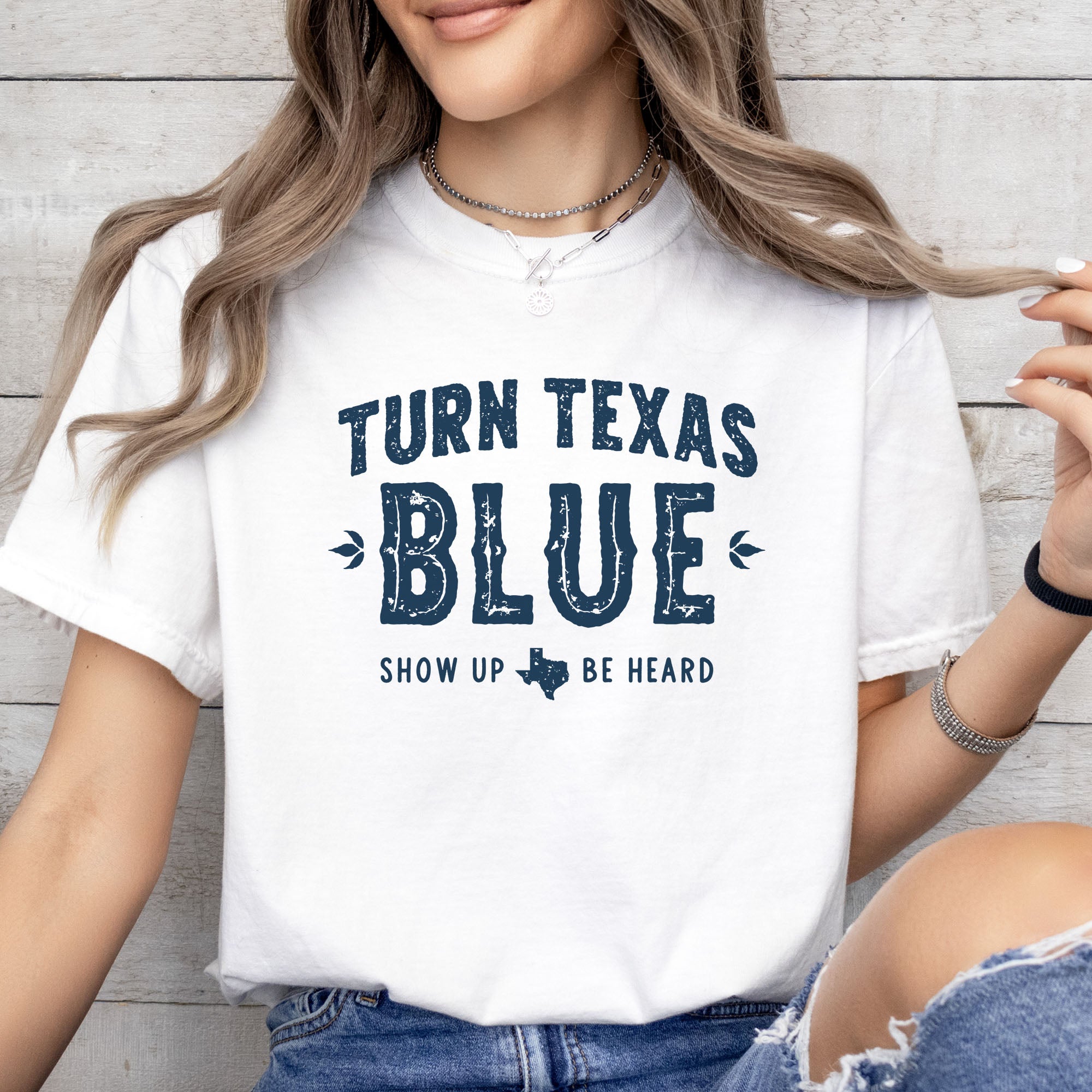 Female model wearing white t-shirt featuring distressed "Turn Texas Blue" text with a small Texas icon and the phrase "Show Up Be Heard."