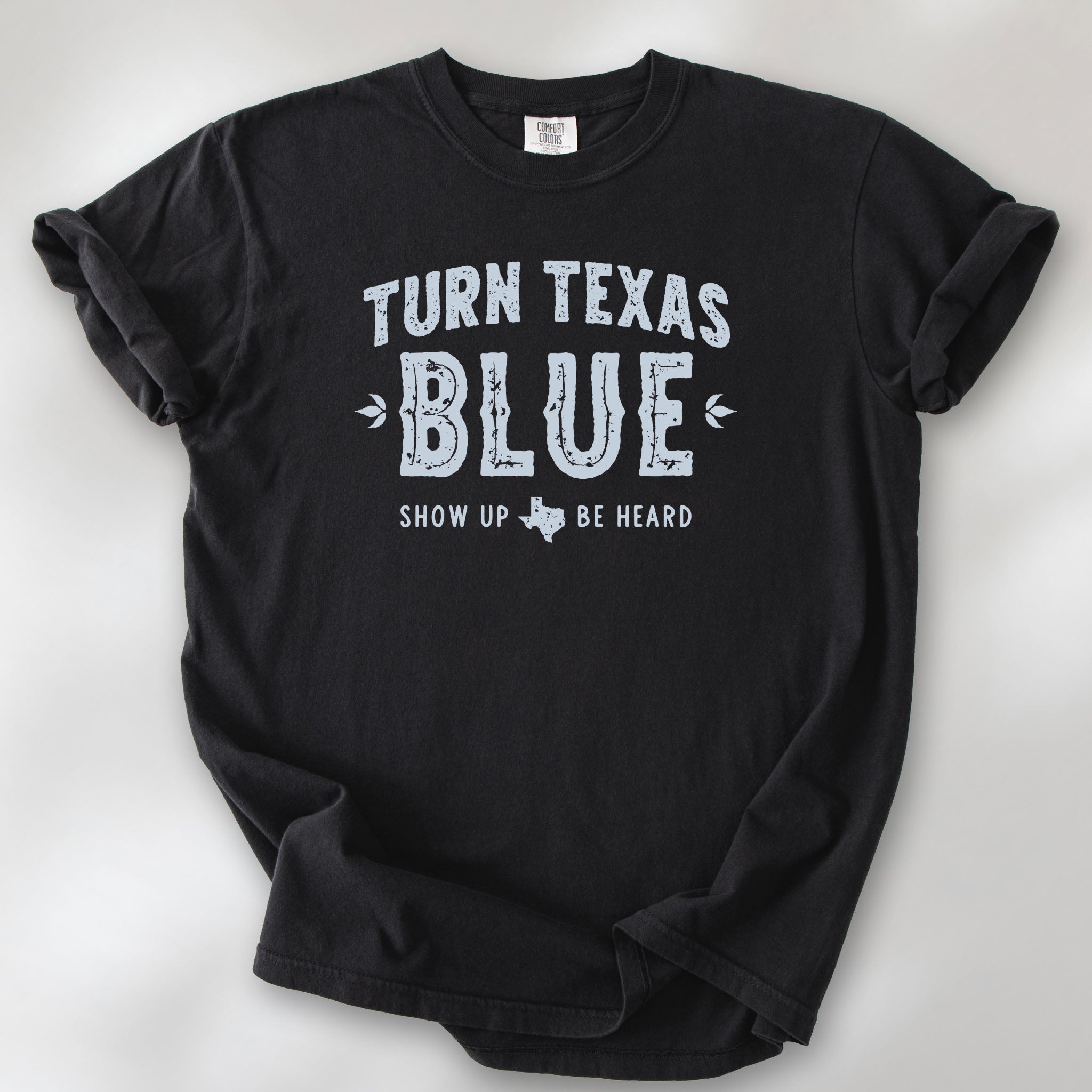 Black t-shirt featuring distressed "Turn Texas Blue" text with a small Texas icon and the phrase "Show Up Be Heard."