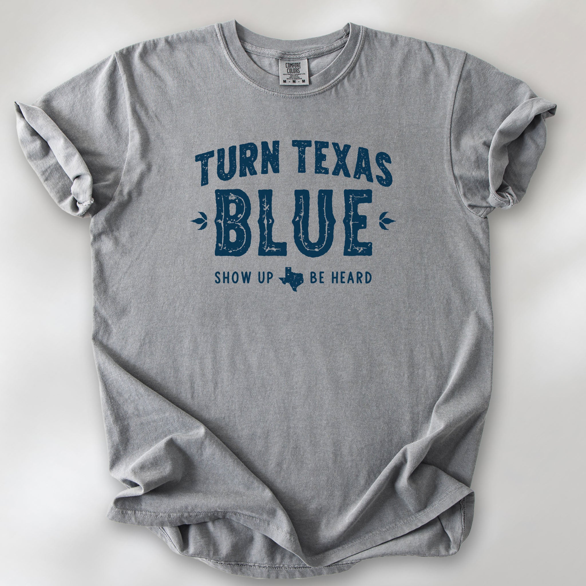Grey t-shirt featuring distressed "Turn Texas Blue" text with a small Texas icon and the phrase "Show Up Be Heard."