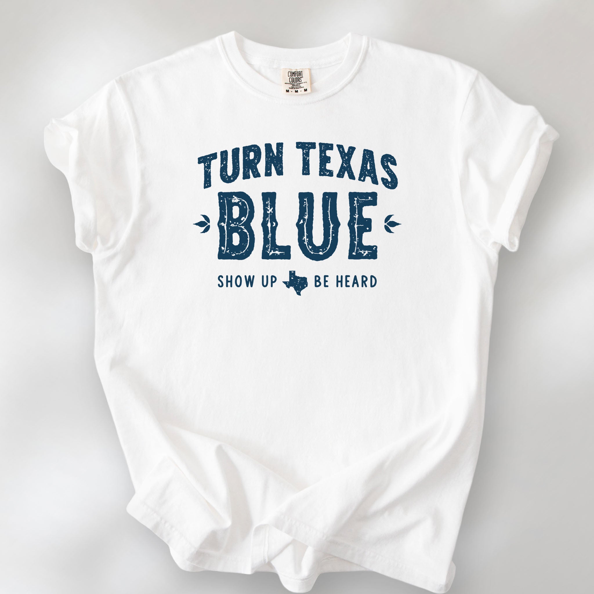 White t-shirt featuring distressed "Turn Texas Blue" text with a small Texas icon and the phrase "Show Up Be Heard."