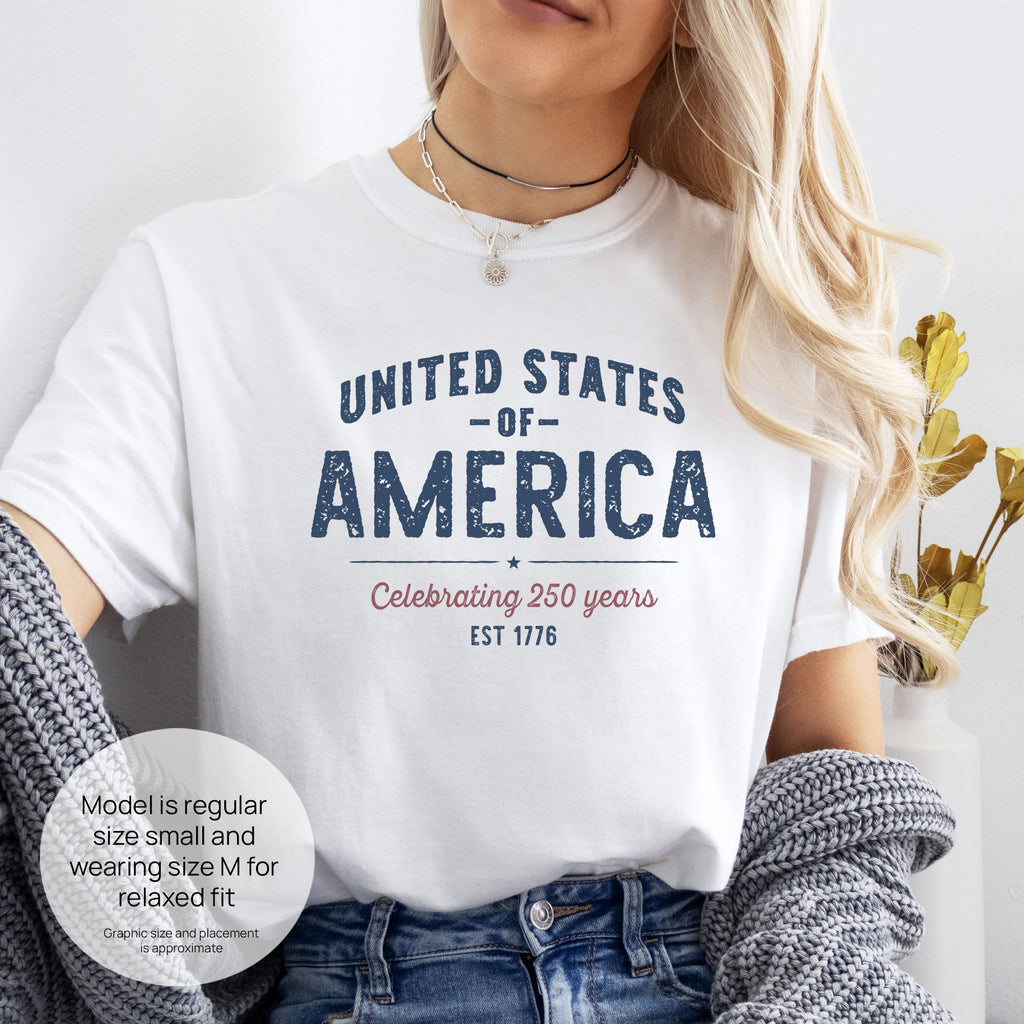 Female model wearing white t-shirt with vintage style text reading “United States of America — Celebrating 250 Years — Est 1776”. Includes size and fit information for the model who is regular size small, and wearing a size M shirt for a relaxed fit.
