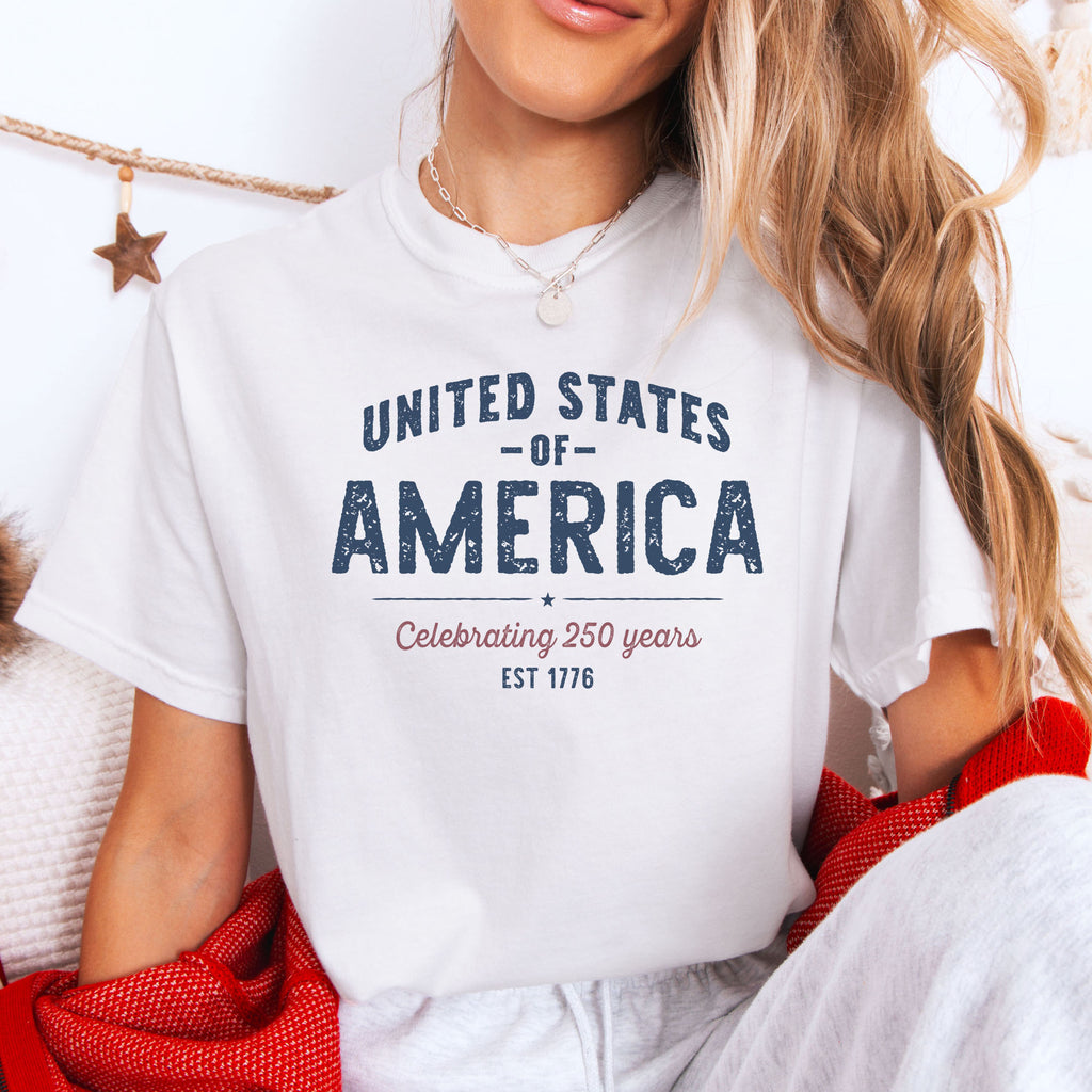Female model wearing white t-shirt with vintage style text reading “United States of America — Celebrating 250 Years — Est 1776”.