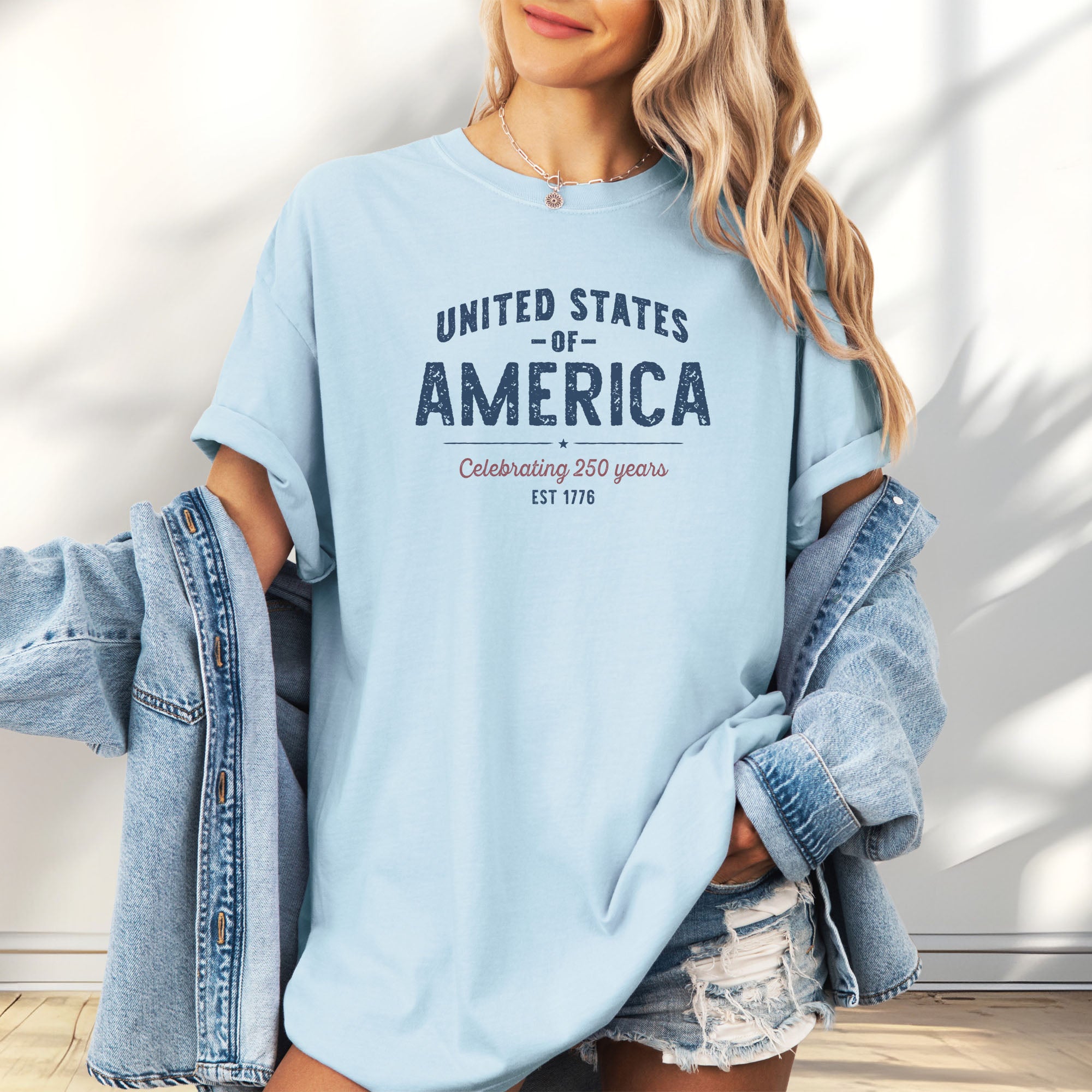 Female model wearing oversized chambray t-shirt with vintage style text reading “United States of America — Celebrating 250 Years — Est 1776”.