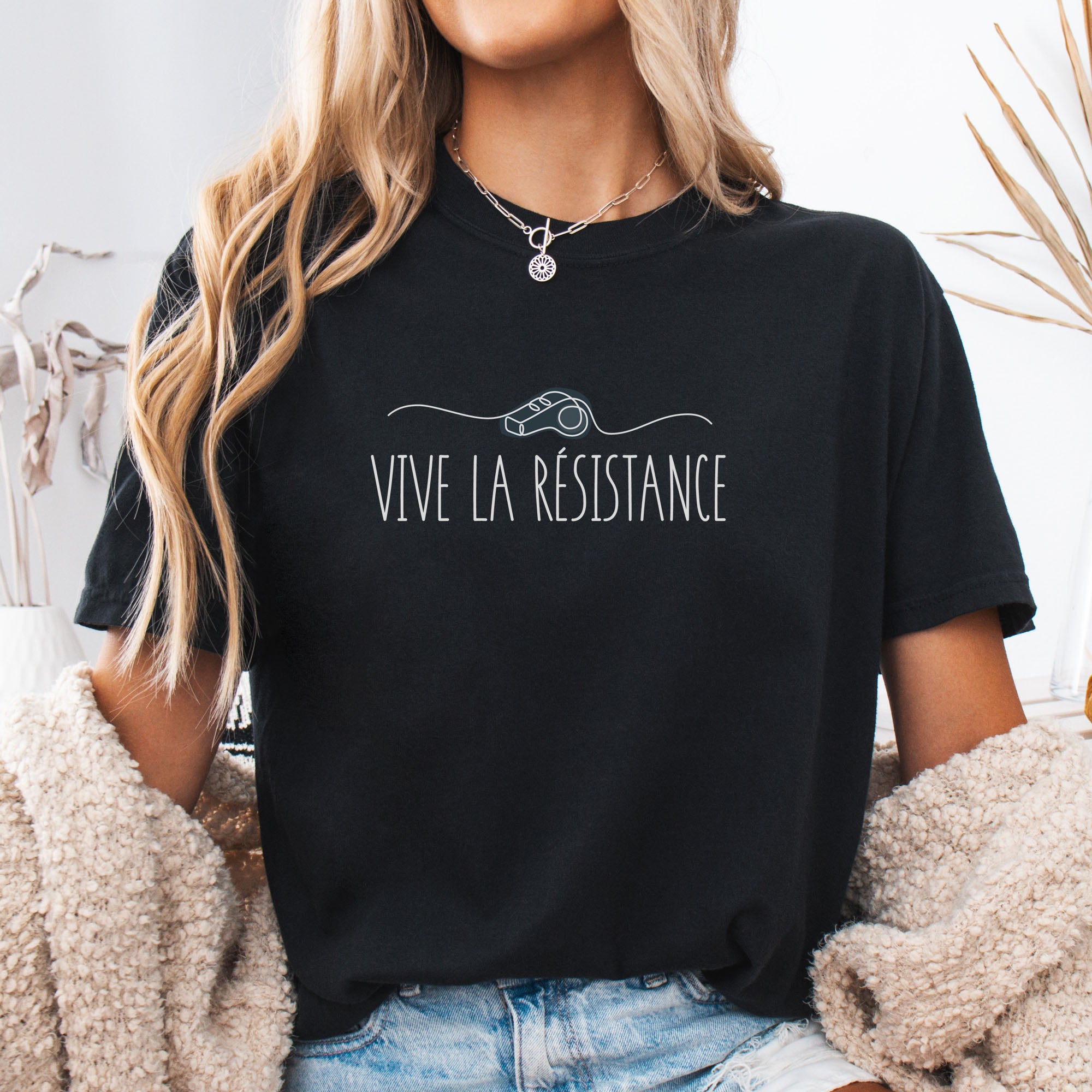 Female model wearing black unisex t-shirt reading “Vive La Resistance” with a whistle illustration referencing community alert systems used to warn neighbors of ICE presence.