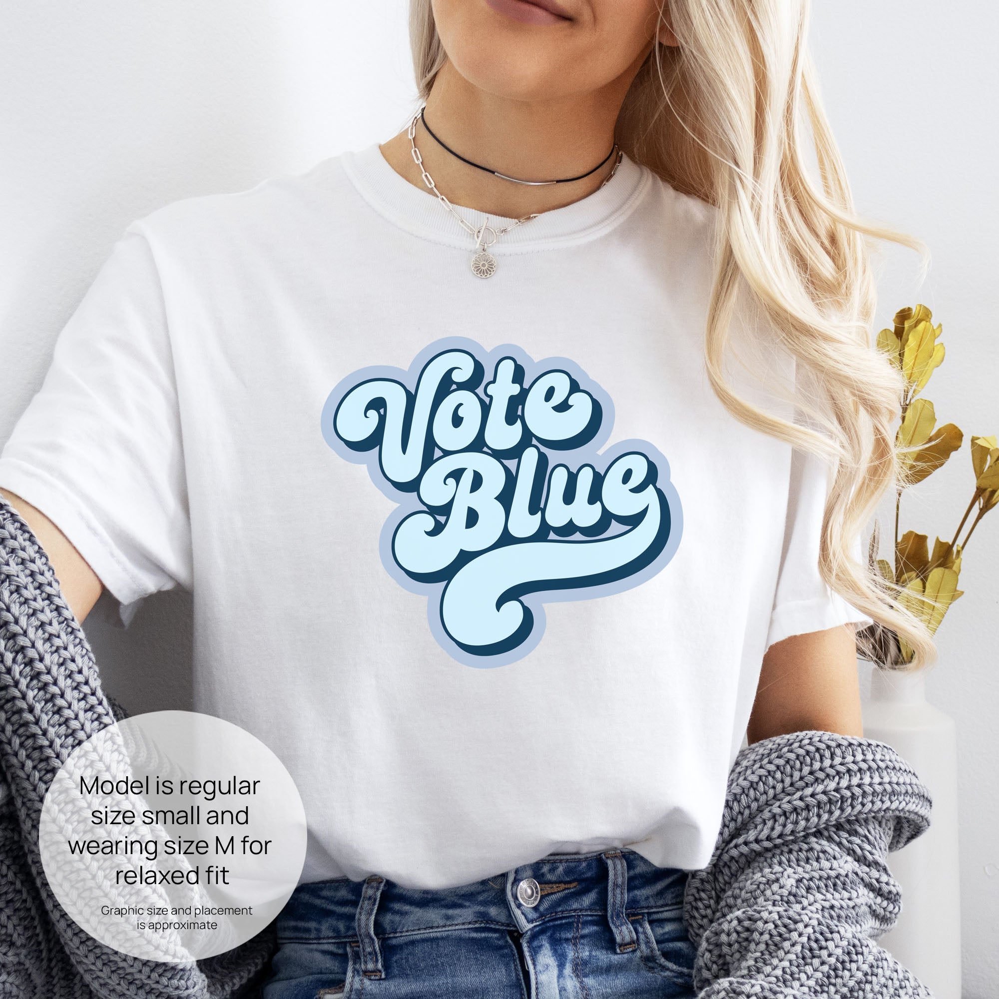 Female model wearing white t-shirt featuring a retro blue "Vote Blue" script graphic. Includes size and fit information for the model who is regular size small, and wearing a size M shirt for a relaxed fit.

