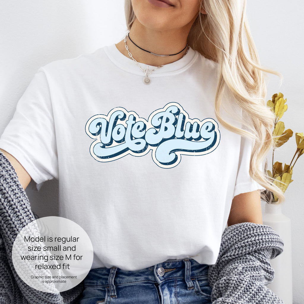 Female model wearing white t-shirt featuring distressed retro script text reading "Vote Blue" in light blue vintage lettering. Includes size and fit information for the model who is regular size small, and wearing a size M shirt for a relaxed fit.
