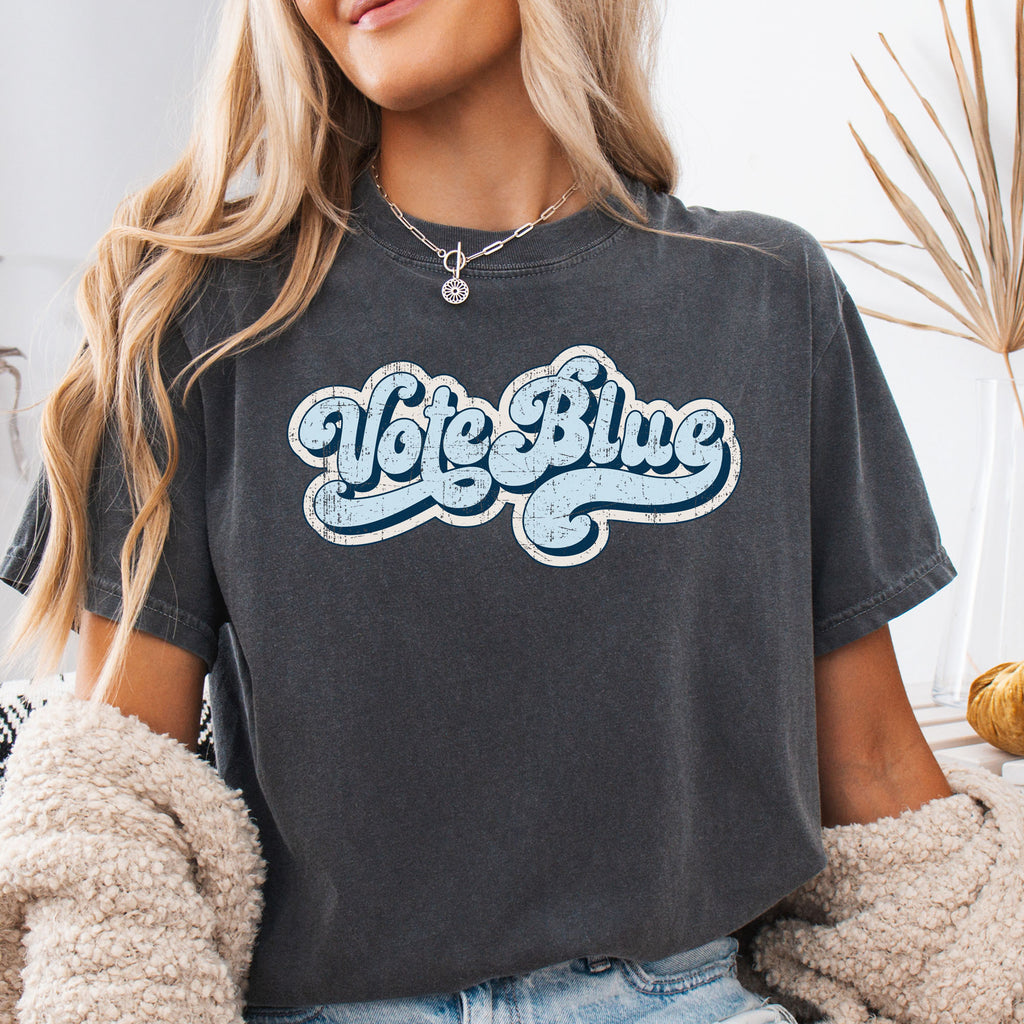 Female model wearing pepper t-shirt featuring distressed retro script text reading "Vote Blue" in light blue vintage lettering.