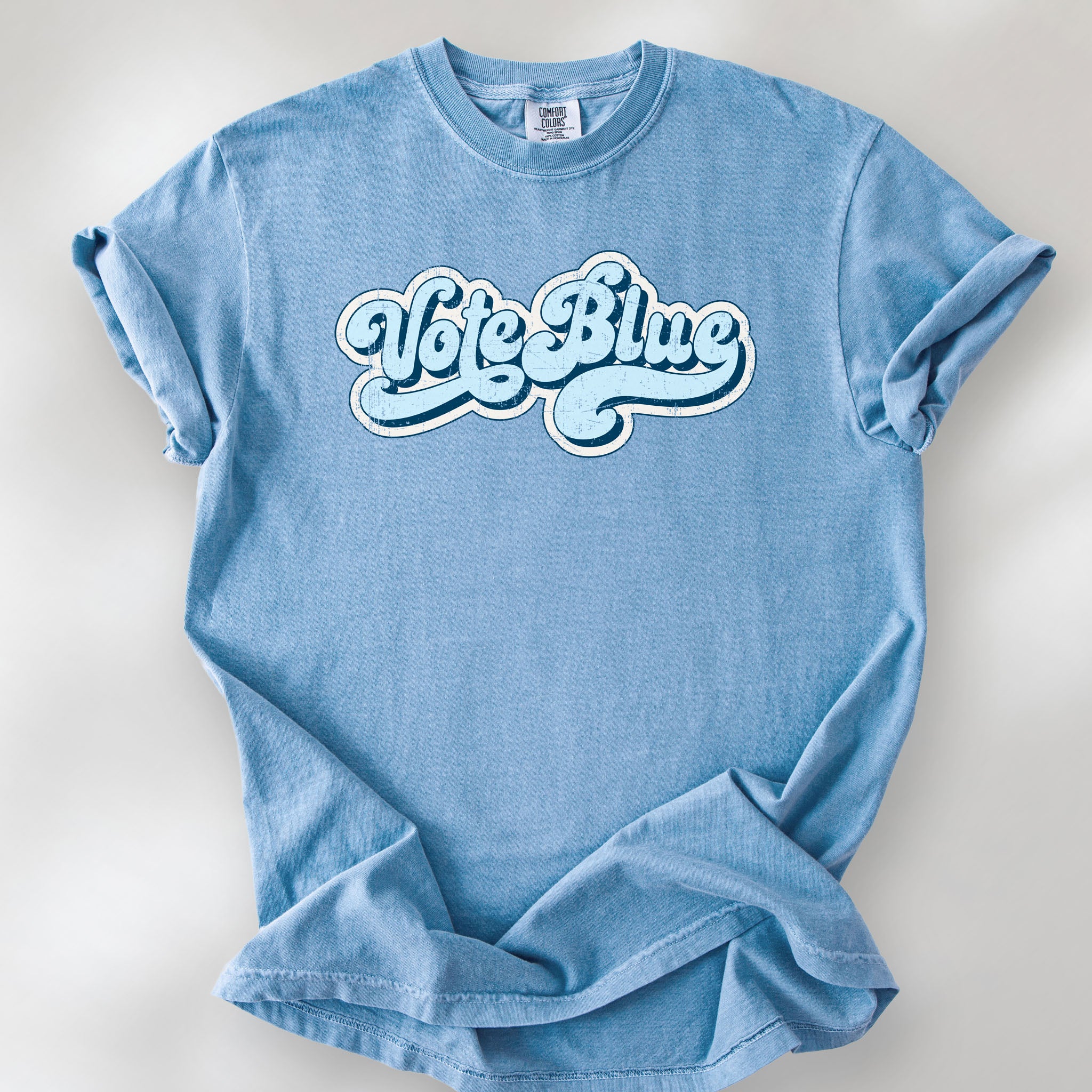 Washed denim t-shirt featuring distressed retro script text reading "Vote Blue" in light blue vintage lettering.