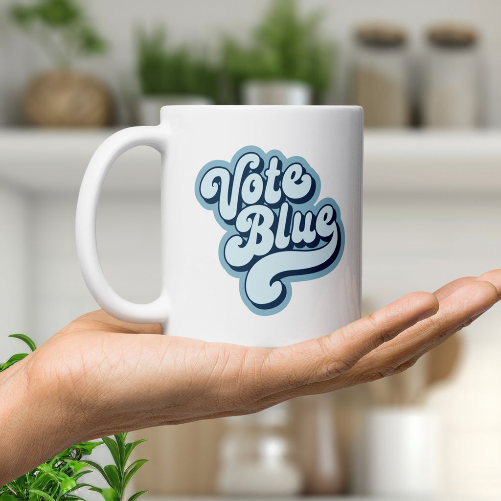 White ceramic coffee mug featuring a retro blue “Vote Blue” script design, displayed in a person's palm in a modern kitchen.