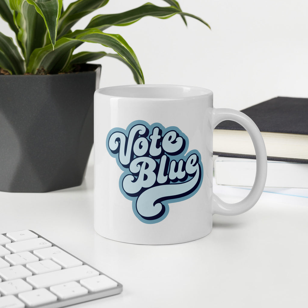 White ceramic coffee mug featuring a retro blue “Vote Blue” script design, sitting on a desk with keyboard, books, and a plant.