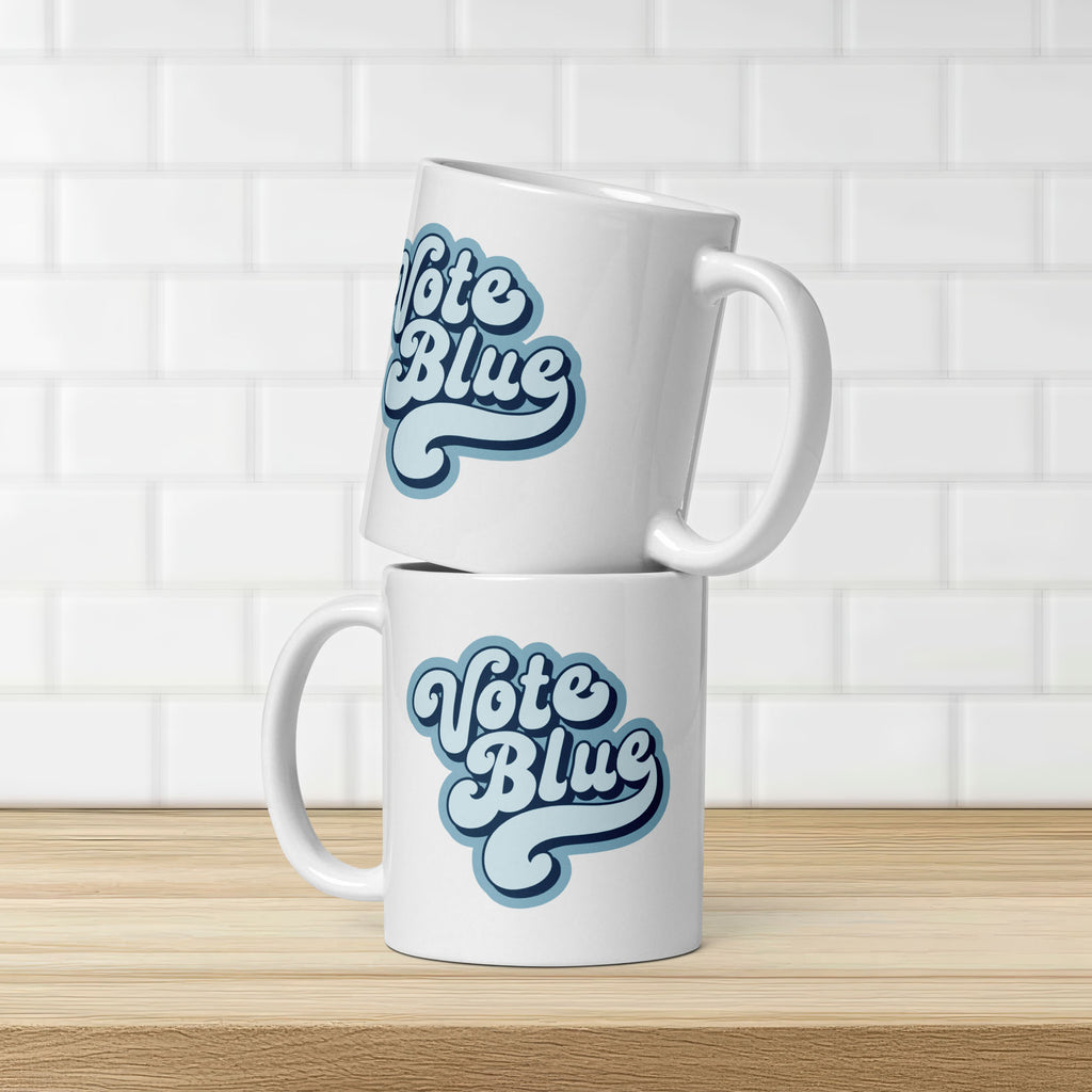 Two white ceramic coffee mug featuring a retro blue “Vote Blue” script design, stacked on a countershop, showing both sides of the mug.