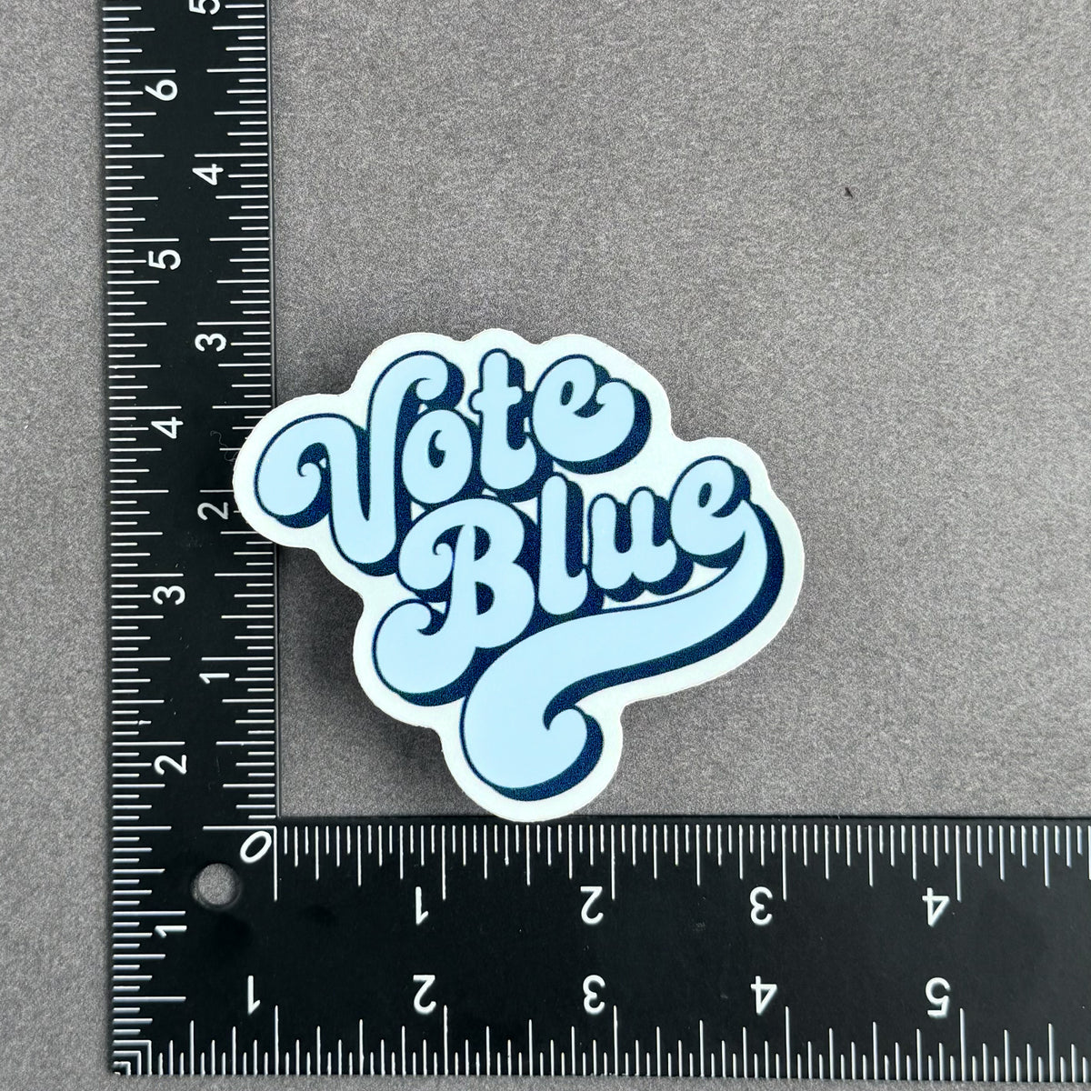 Die-cut Vote Blue vinyl sticker with retro blue typography pictured with a ruler to show approximate 3 inch by 3 inch size.