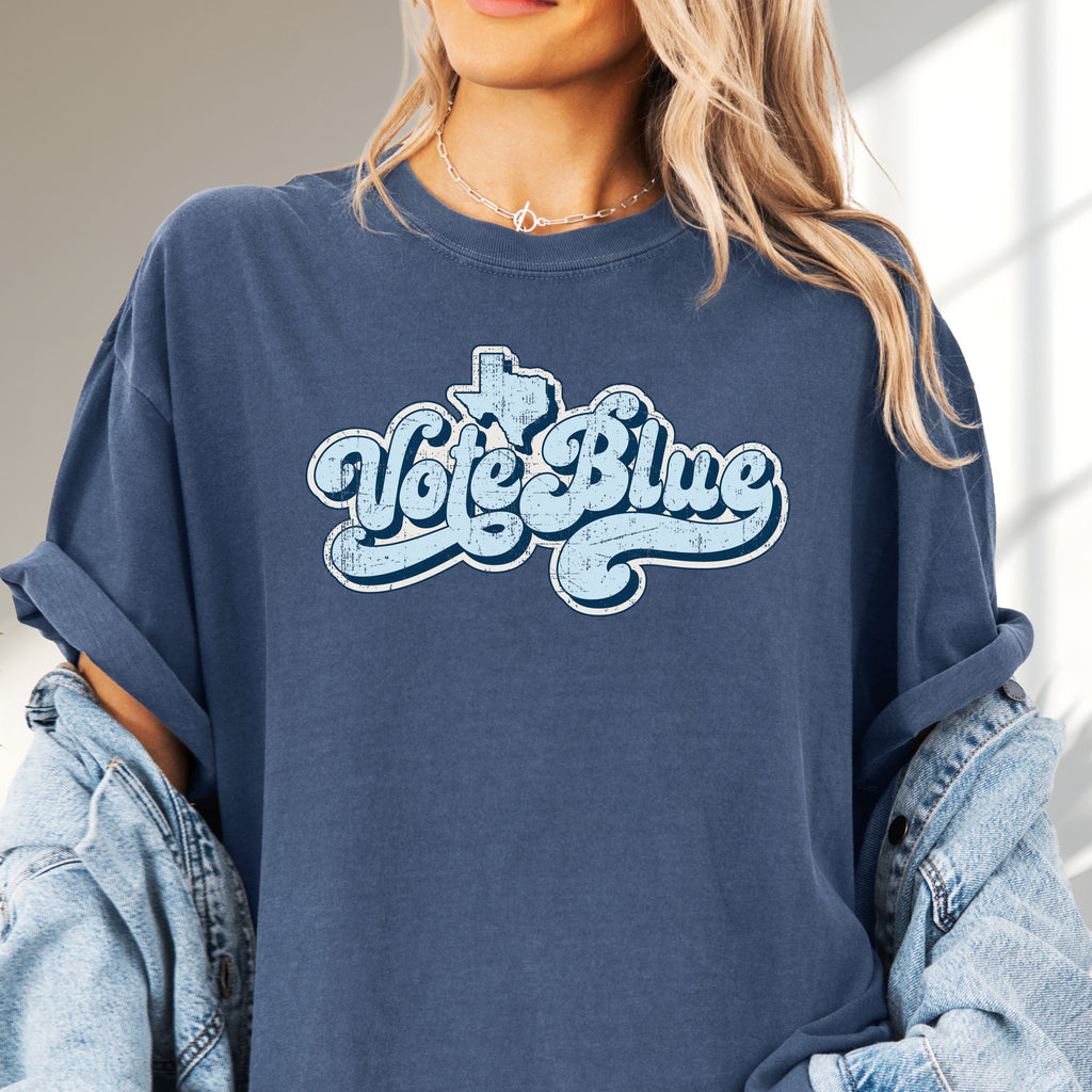 Female model wearing denim t-shirt featuring distressed retro "Vote Blue" script lettering with the outline of Texas above the design.