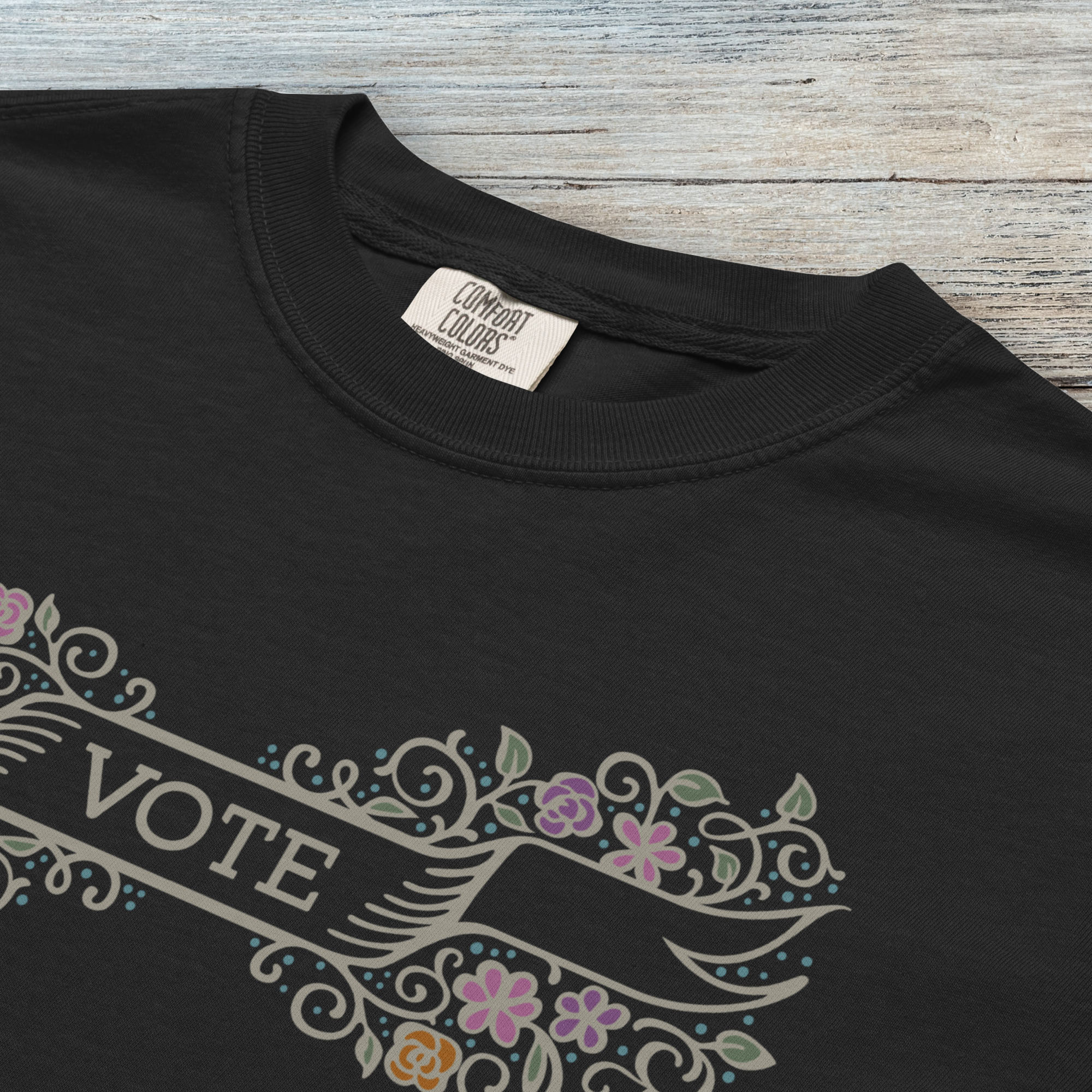 Closeup detail of black t-shirt with vintage style floral illustration and a banner reading “vote” in the center.