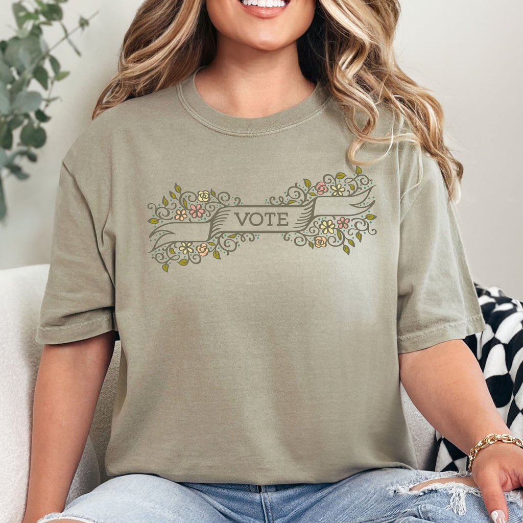 Female model wearing khaki t-shirt with vintage style floral illustration and a banner reading “vote” in the center.