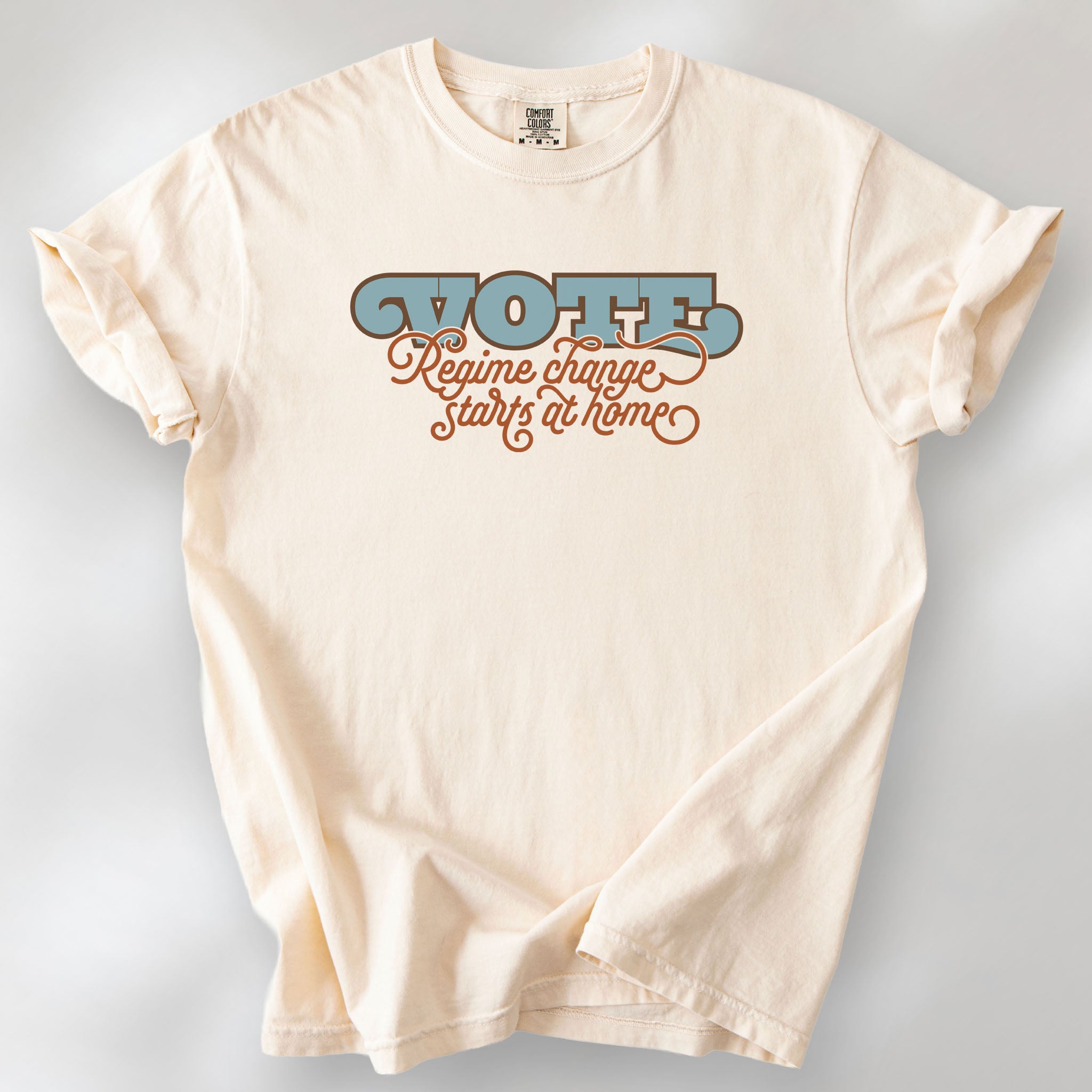 Ivory t-shirt with retro text reading “Vote: Regime Change Starts at Home,” promoting voting and political accountability.