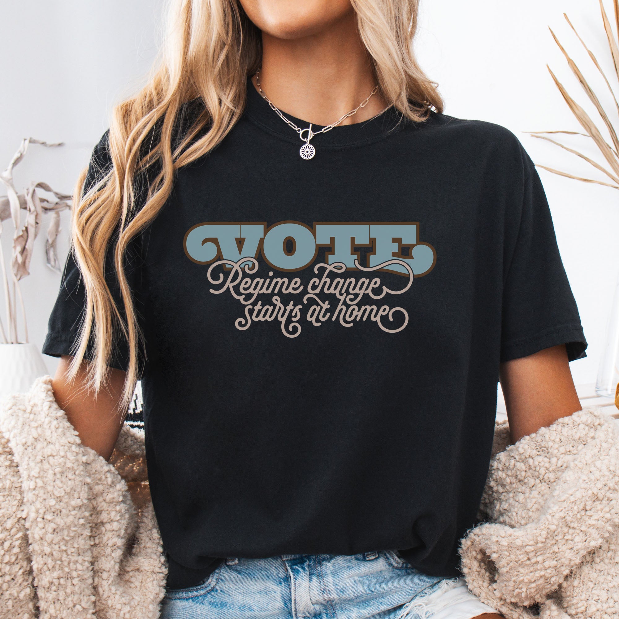 Female model wearing black t-shirt with retro text reading “Vote: Regime Change Starts at Home,” promoting voting and political accountability.