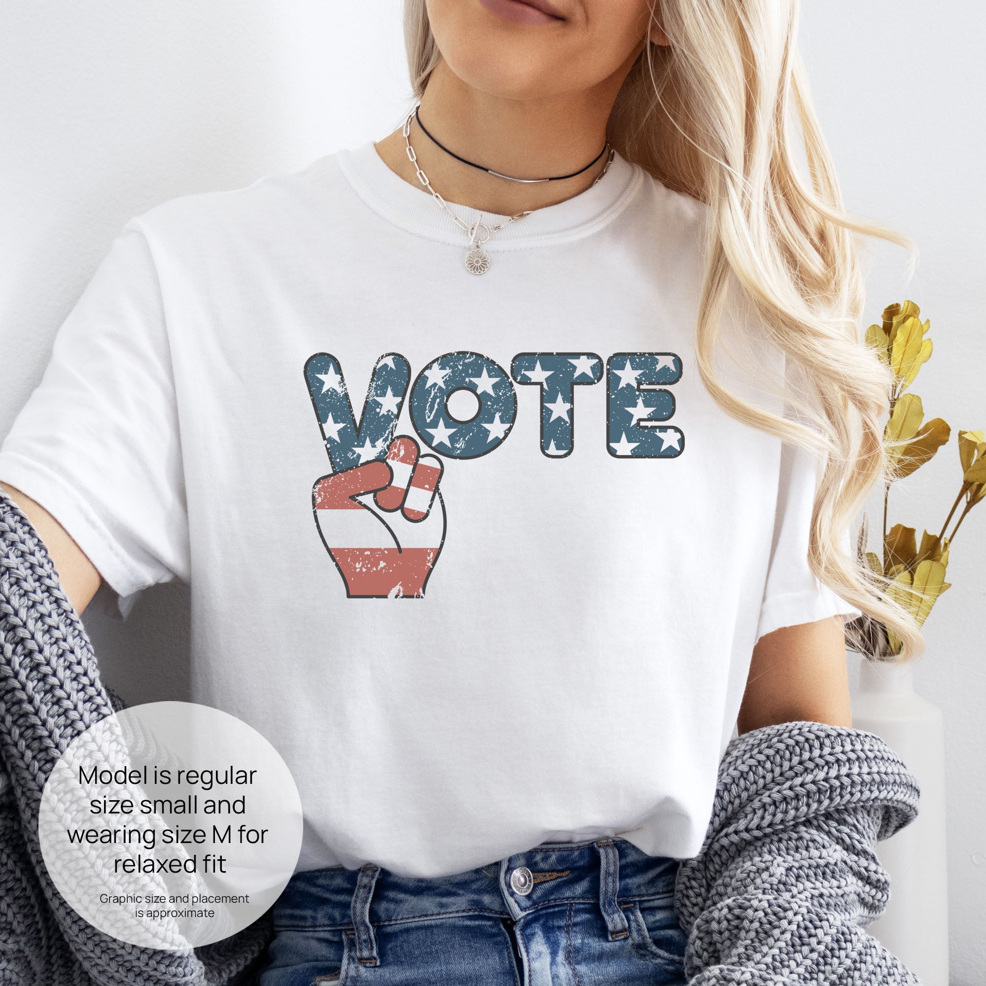 Female model wearing white t-shirt featuring a distressed retro "VOTE" graphic with blue stars and a vintage red and blue hand making a peace sign. Includes size and fit information for the model who is regular size small, and wearing a size M shirt for a relaxed fit.
