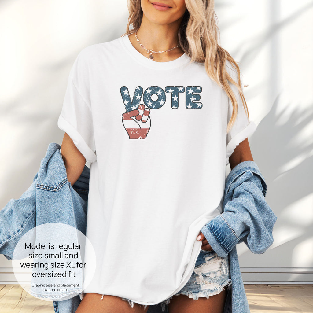 Female model wearing oversized white t-shirt featuring a distressed retro "VOTE" graphic with blue stars and a vintage red and blue hand making a peace sign. Includes size and fit information for the model who is regular size small, and wearing a size XL shirt for an oversized fit.
