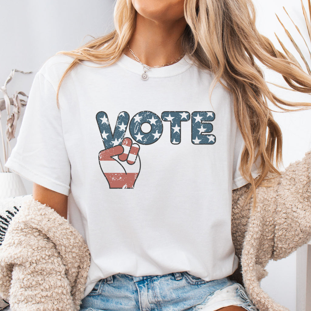 Female model wearing white t-shirt featuring a distressed retro "VOTE" graphic with blue stars and a vintage red and blue hand making a peace sign.