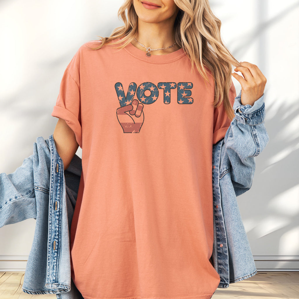 Female model wearing oversized terracotta t-shirt featuring a distressed retro "VOTE" graphic with blue stars and a vintage red and blue hand making a peace sign.