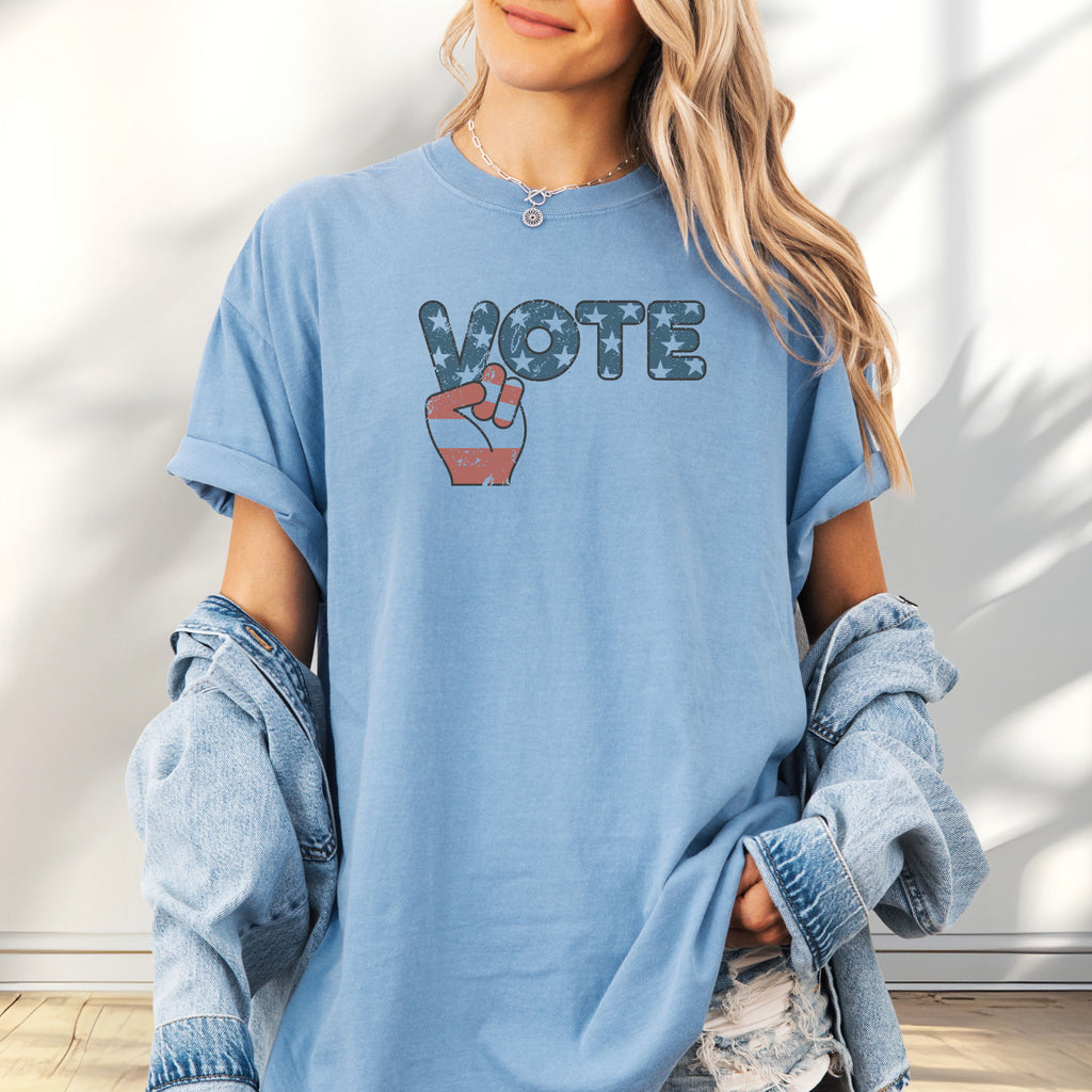 Female model wearing oversized washed denim t-shirt featuring a distressed retro "VOTE" graphic with blue stars and a vintage red and blue hand making a peace sign.
