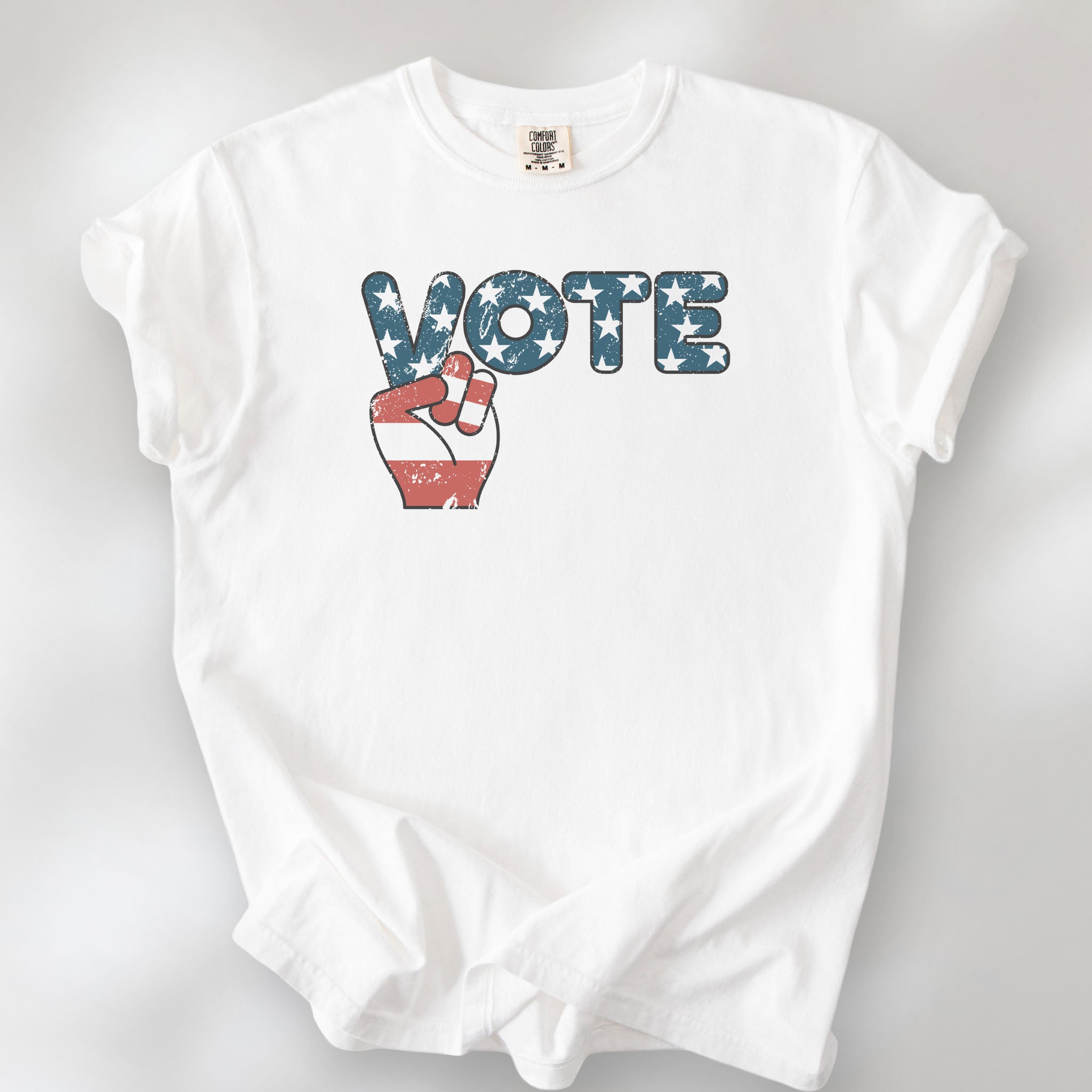 White t-shirt featuring a distressed retro "VOTE" graphic with blue stars and a vintage red and blue hand making a peace sign.