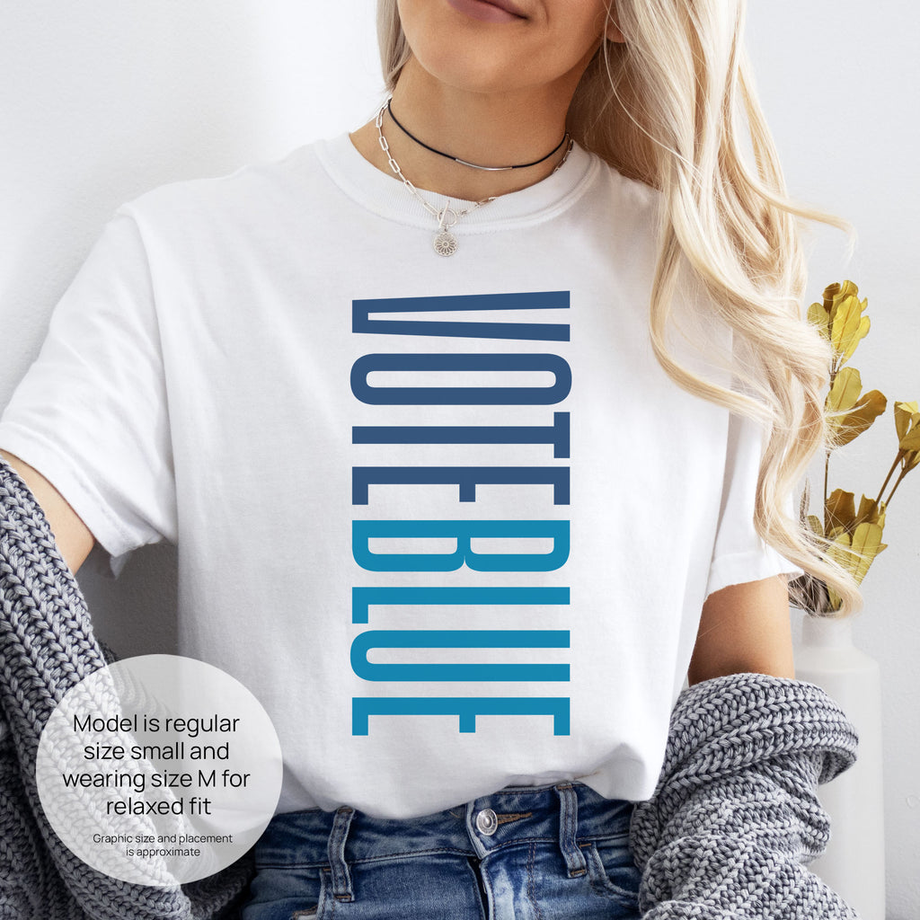 Female model wearing white t-shirt featuring the words "VOTE BLUE" stacked vertically in bold minimalist blue typography. Includes size and fit information for the model who is regular size small, and wearing a size M shirt for a relaxed fit.
