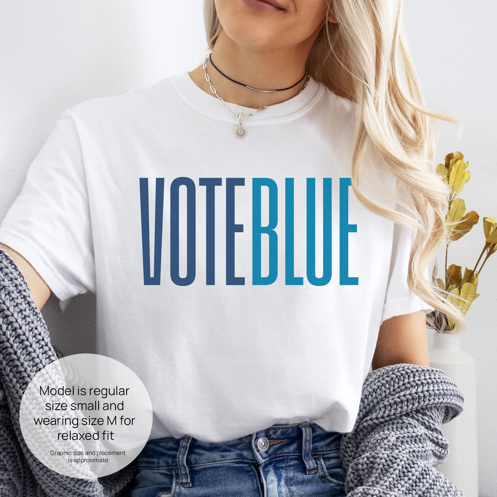 Female model wearing white t-shirt featuring the words "VOTE BLUE" in bold horizontal blue geometric typography with a clean minimalist design. Includes size and fit information for the model who is regular size small, and wearing a size M shirt for a relaxed fit.
