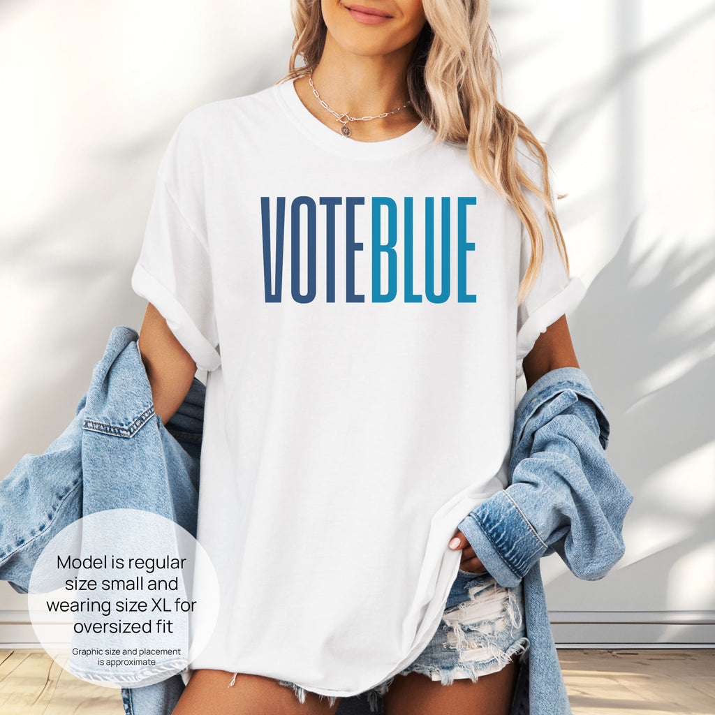Female model wearing oversized white t-shirt featuring the words "VOTE BLUE" in bold horizontal blue geometric typography with a clean minimalist design. Includes size and fit information for the model who is regular size small, and wearing a size XL shirt for an oversized fit.
