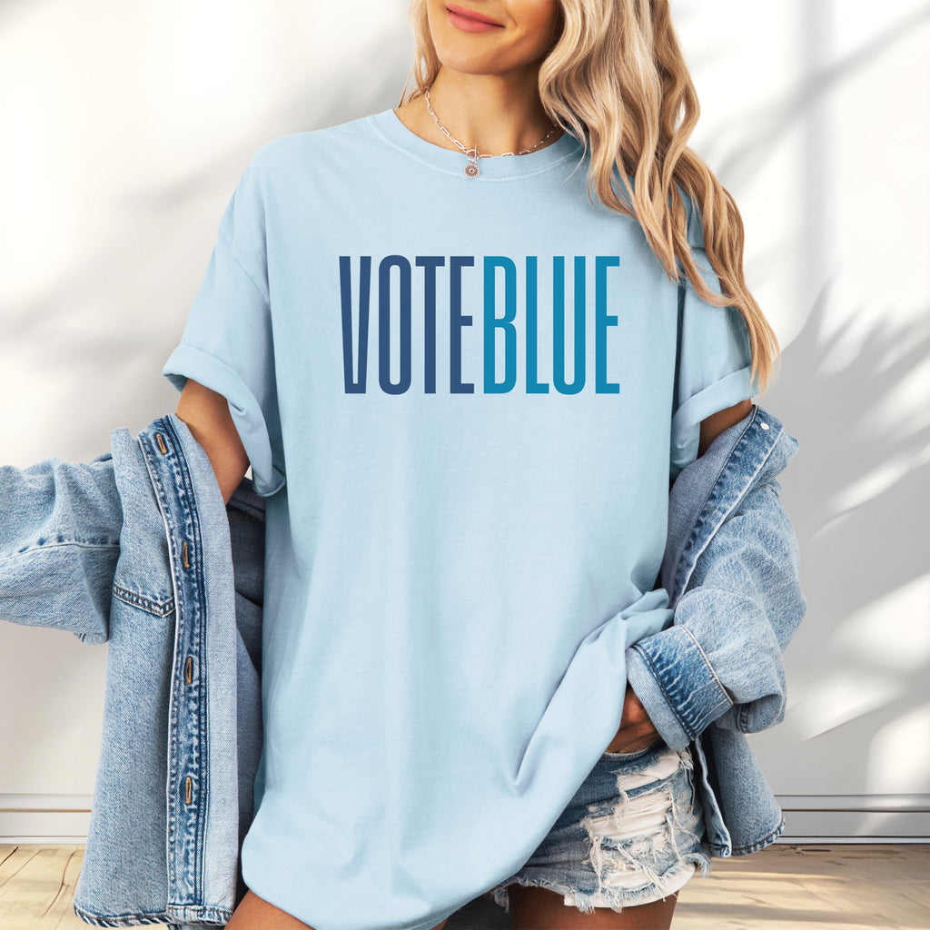 Female model wearing oversized chambray t-shirt featuring the words "VOTE BLUE" in bold horizontal blue geometric typography with a clean minimalist design.