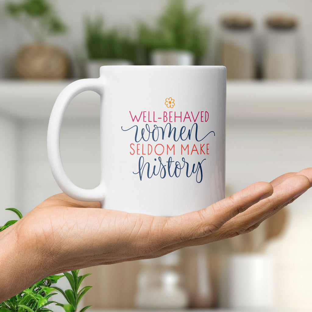 White mug with motivational quote that reads "well-behaved women seldom make history" held in the palm of a hand.