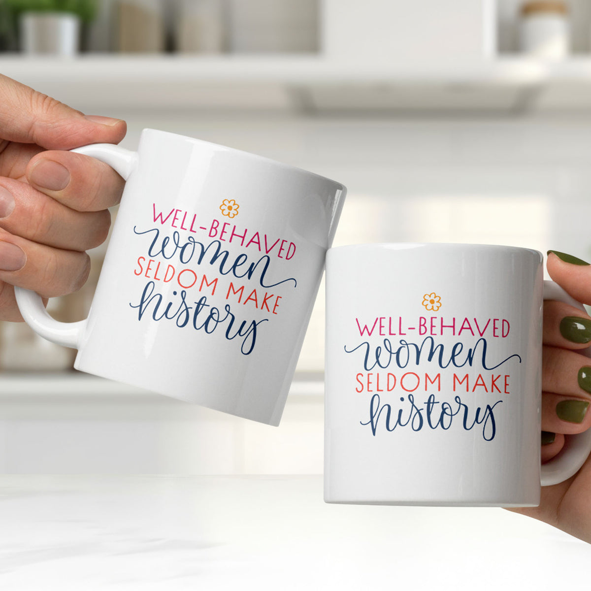 Two white ceramic mugs with motivational quote that reads "well-behaved women seldom make history" held by two people clinking them together in a bright white kitchen.