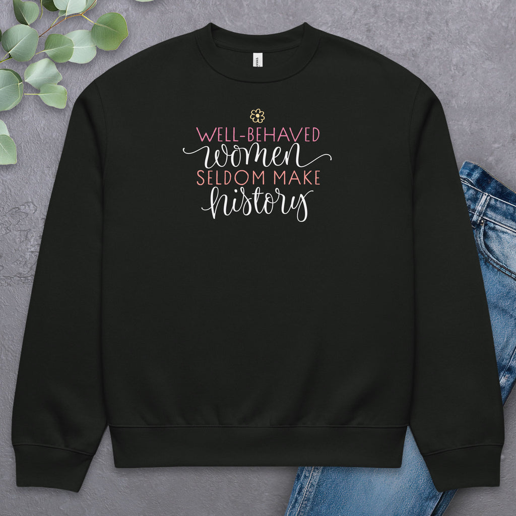 Black crewneck sweatshirt with stylized text graphic that reads "well-behaved women seldom make history",  laying flat with a pair of jeans.
