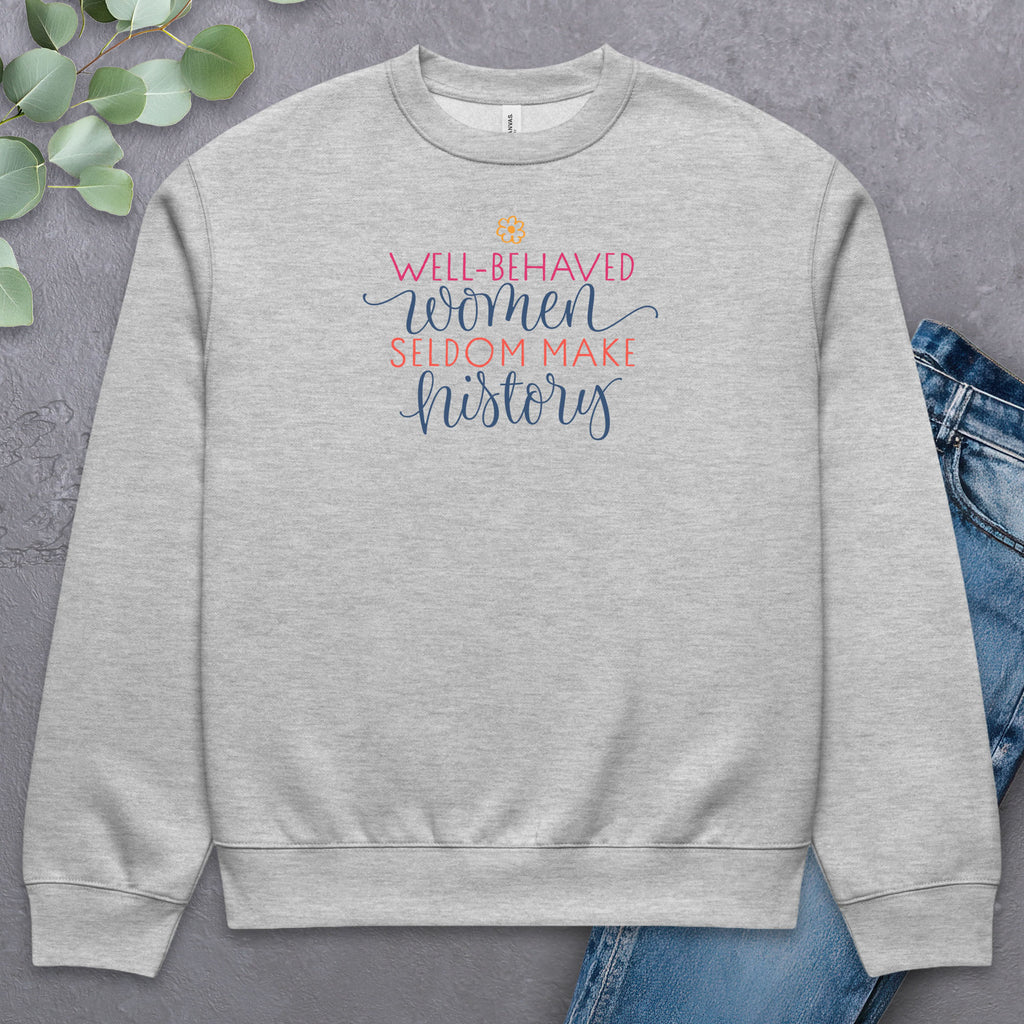 Athletic heather grey crewneck sweatshirt with stylized text graphic that reads "well-behaved women seldom make history",  laying flat with a pair of jeans.