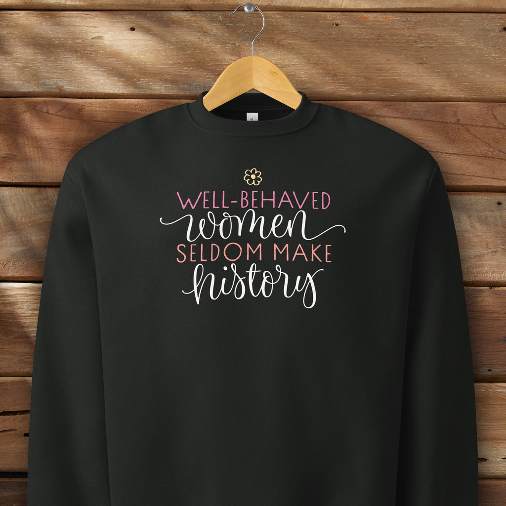 Black crewneck sweatshirt with stylized text graphic that reads "well-behaved women seldom make history" hanging on a hanger.