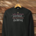 Black crewneck sweatshirt with stylized text graphic that reads "well-behaved women seldom make history" hanging on a hanger.
