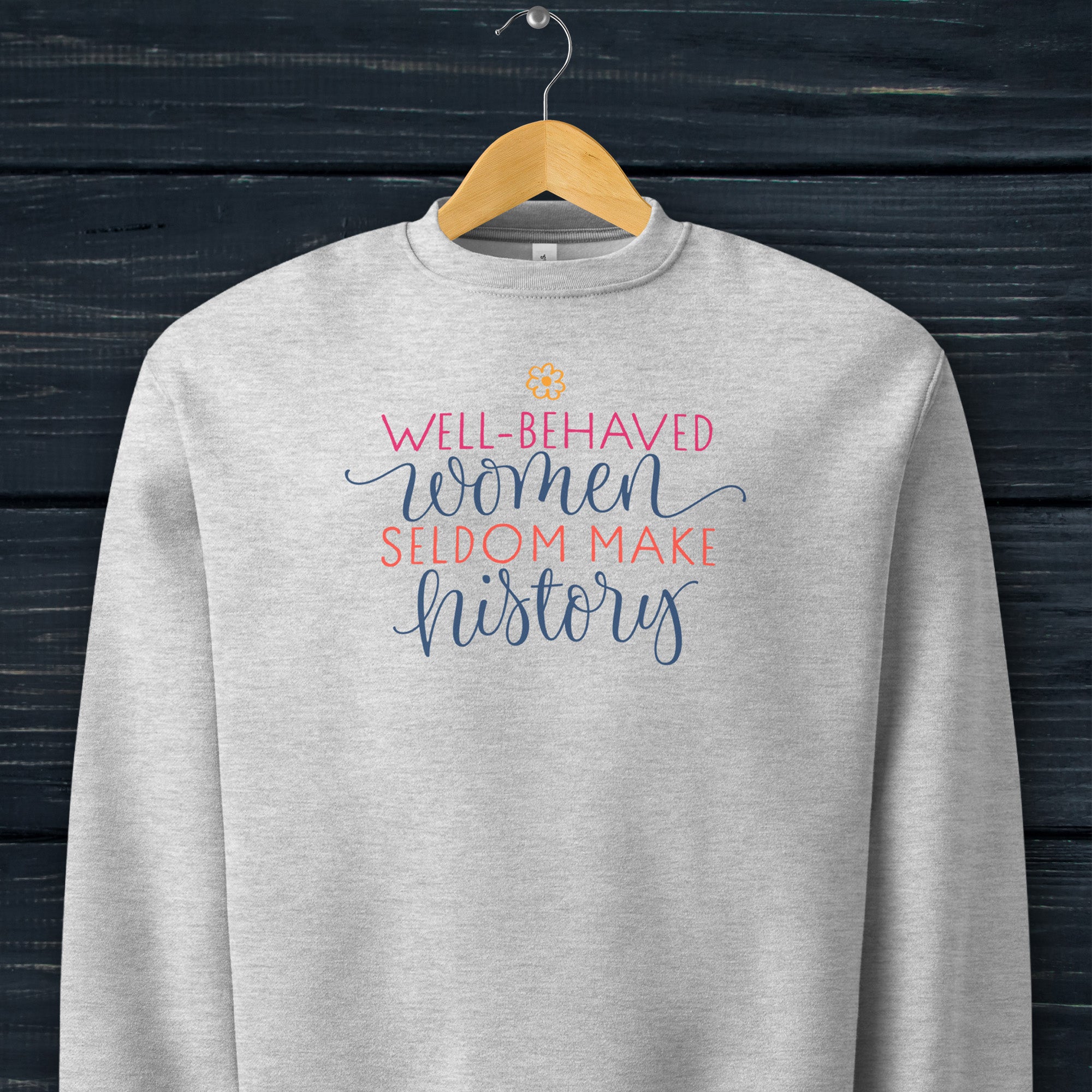 Athletic heather grey crewneck sweatshirt with stylized text graphic that reads "well-behaved women seldom make history" hanging on a hanger.