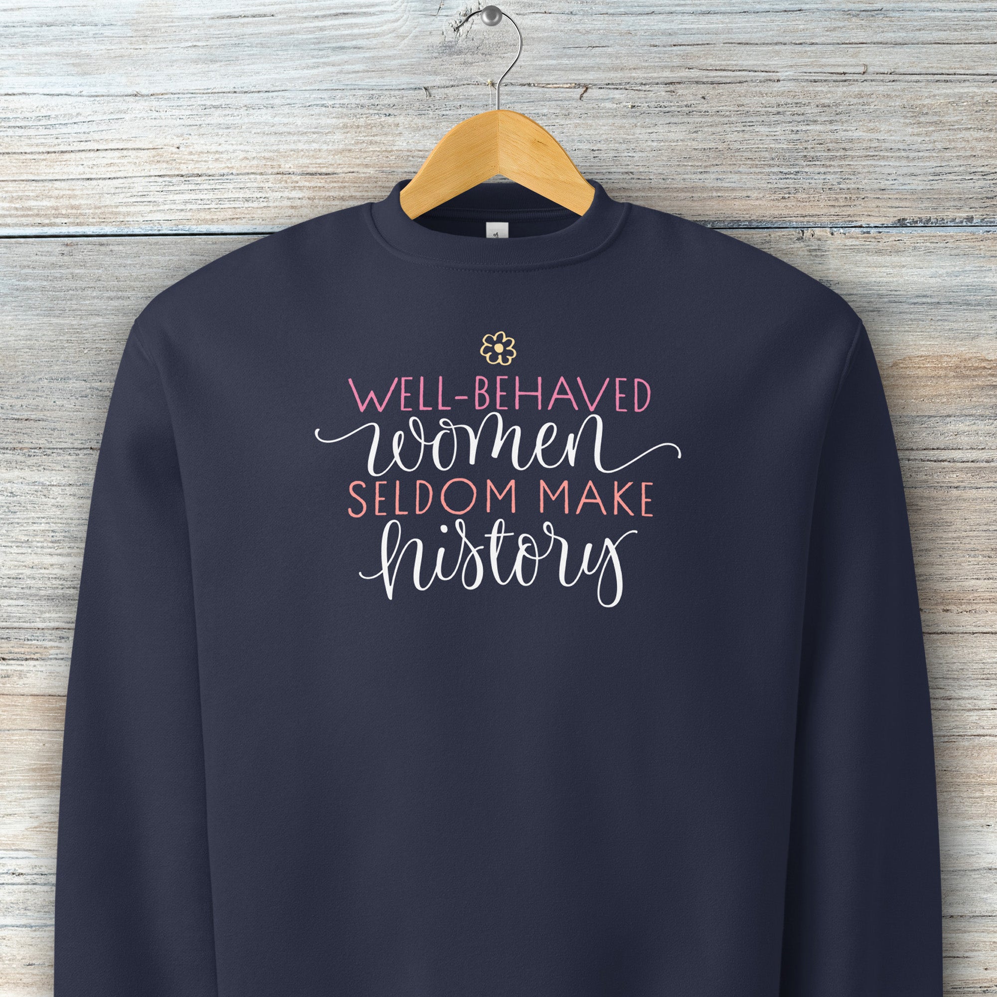 Navy crewneck sweatshirt with stylized text graphic that reads "well-behaved women seldom make history" hanging on a hanger.