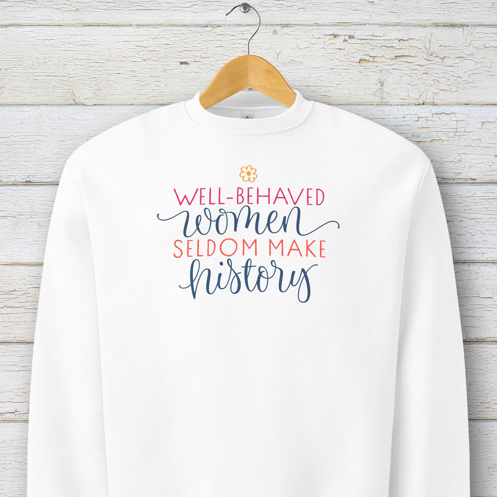 White crewneck sweatshirt with stylized text graphic that reads "well-behaved women seldom make history" hanging on a hanger.