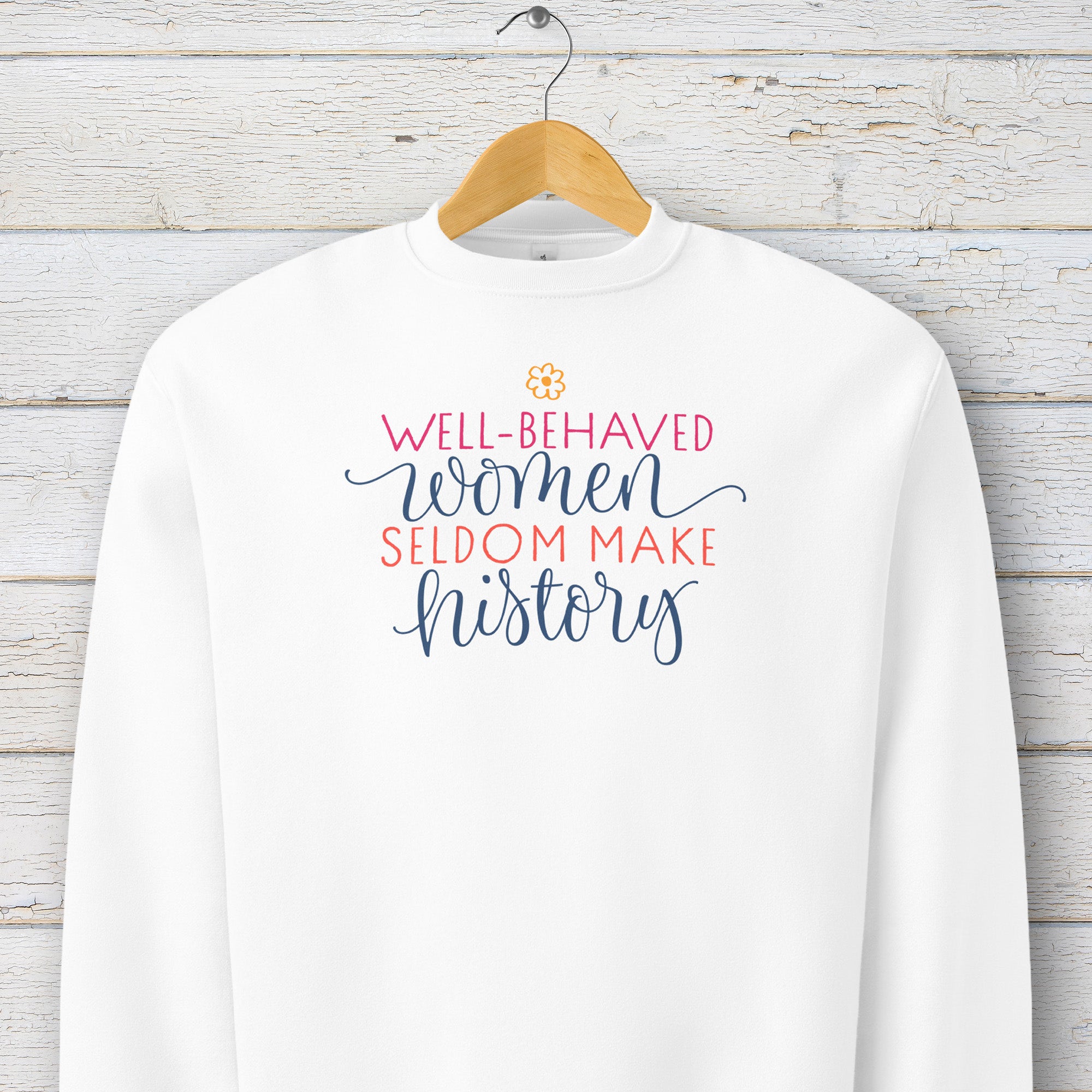 White crewneck sweatshirt with stylized text graphic that reads "well-behaved women seldom make history" hanging on a hanger.