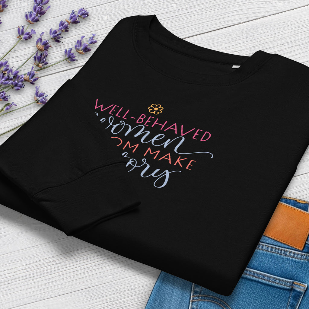 Folded black crewneck sweatshirt with stylized text graphic that reads "well-behaved women seldom make history", styled with a pair of jeans and purple flowers.