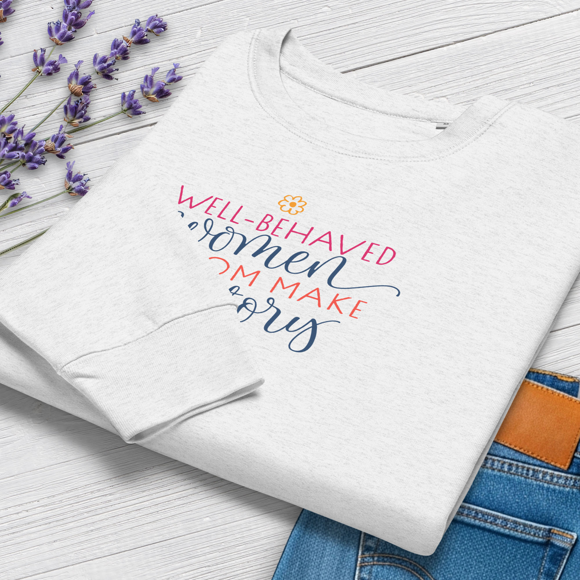 Folded grey crewneck sweatshirt with stylized text graphic that reads "well-behaved women seldom make history", styled with a pair of jeans and purple flowers.