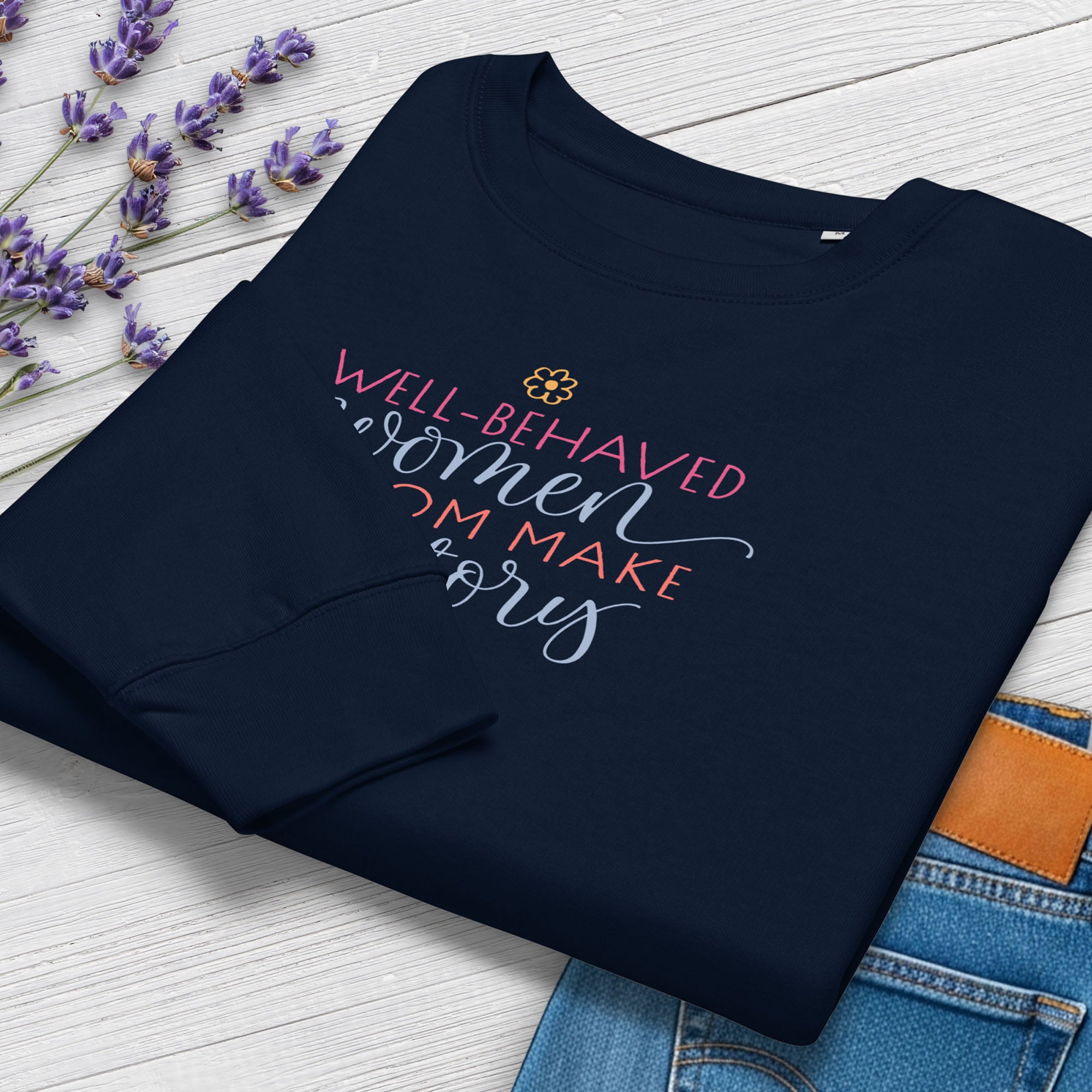 Folded blue crewneck sweatshirt with stylized text graphic that reads "well-behaved women seldom make history", styled with a pair of jeans and purple flowers.