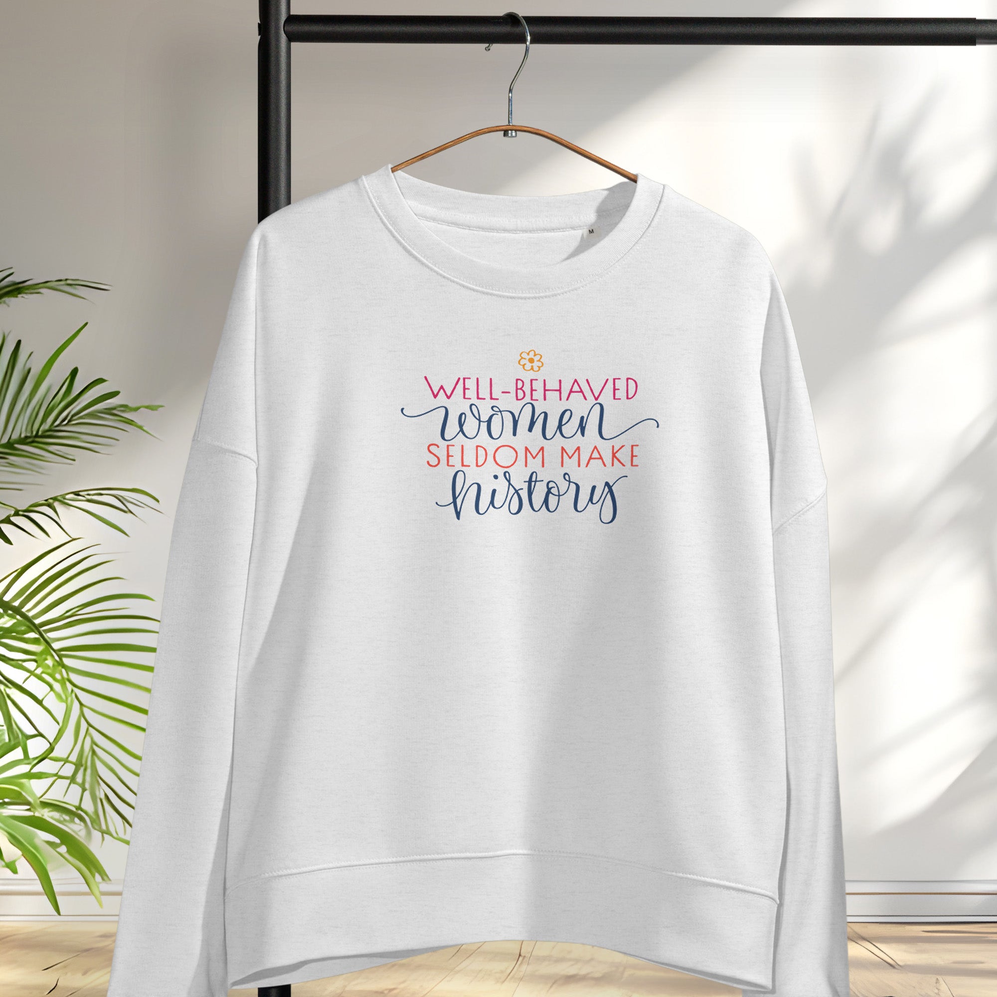 Gret crewneck sweatshirt with stylized text graphic that reads "well-behaved women seldom make history" hanging on a hanger.